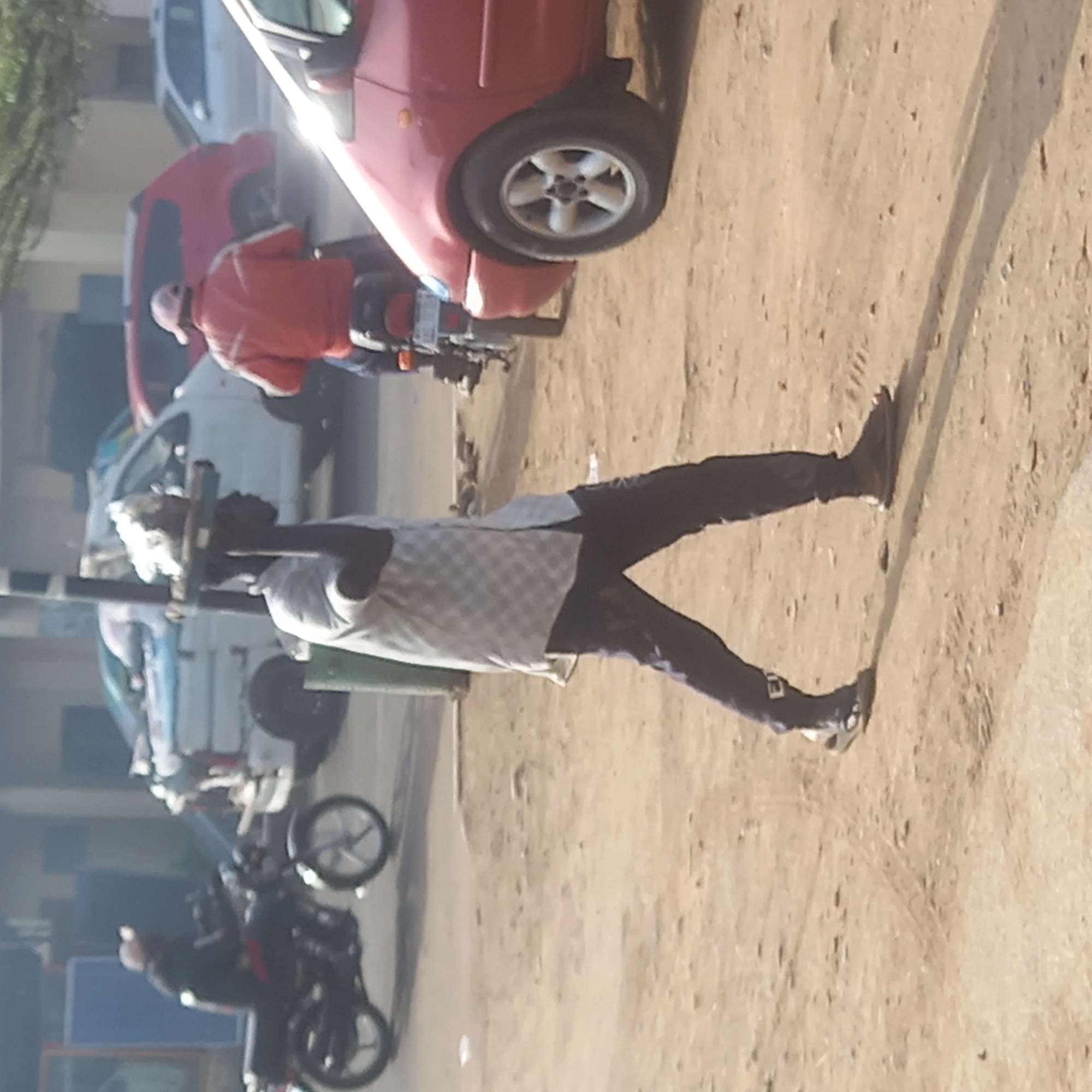 This outdoor image, likely taken in Dadi Riba, Nigeria, depicts a busy street scene under bright daylight, suggested by the clear visibility and distinct, long shadows cast on the unpaved, sandy ground.
In the foreground, a person is seen walking towards the right side of the frame, carrying a long, dark pole over their shoulder. They are dressed in a light-colored, possibly checkered, shirt, dark trousers, and sandals. A white cloth or bag is visible at the end of the pole near their head.
To their left, in the middle ground, another individual sits on a motorcycle. This person is wearing an orange or red jacket and a light-colored cap or helmet. Behind this motorcycle, a red car is parked, partially obscuring a white car further back. Another dark-colored motorcycle with a rider is visible further in the background, indicating ongoing street activity. Buildings and sparse trees line the background. There is no visible text on any signs or vehicles.
