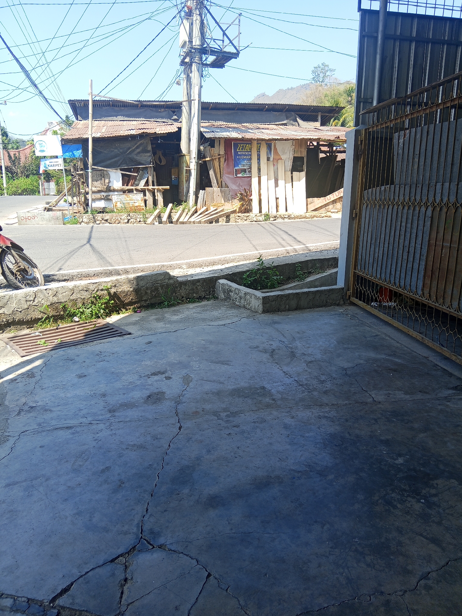 The image shows a street scene in Makale, Indonesia, under a bright, clear sky. In the foreground, a cracked concrete surface transitions to a paved road. A red motorcycle is partially visible on the left, parked near a drainage grate. Beyond the road, a collection of structures with corrugated metal roofs and wooden supports are arranged. One of these buildings displays a sign that reads "ZETHA" and "KARPET", indicating a business. The structures appear somewhat dilapidated. Overhead, a dense network of electrical wires crisscrosses the sky. To the right of the frame, a tall, rusted metal gate partially obscures a dark interior space. In the background, a hilly, tree-covered landscape is visible.