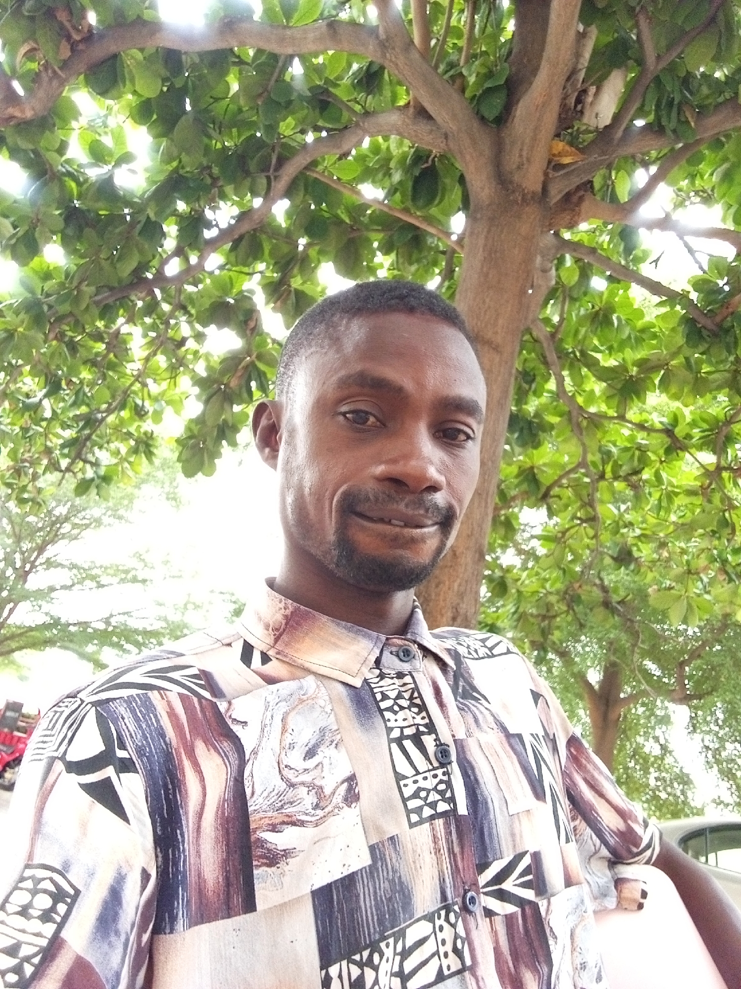 A man stands beneath a tree in Lokoja, Nigeria. The tree is tall with a thick trunk and lush green leaves that create a canopy overhead. The man has short black hair, dark eyes, and a beard. He is wearing a patterned button-down shirt. The background includes other trees and hints of buildings.