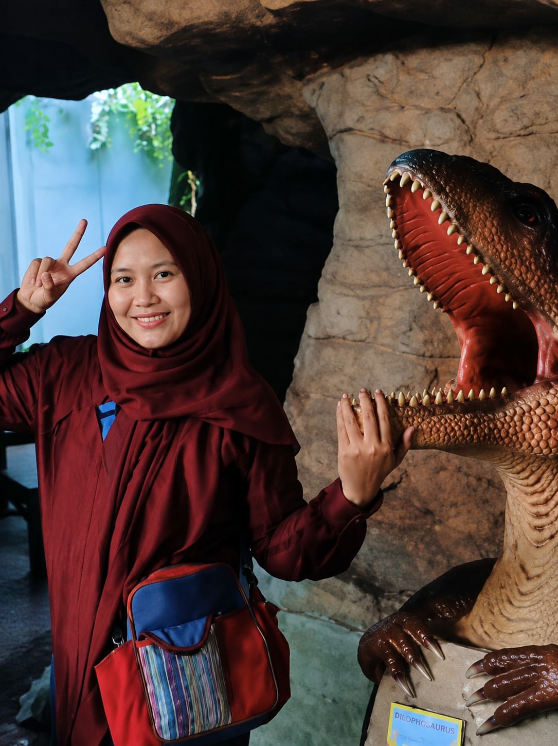 In a cave-like environment at the Geological Museum of Bandar Baru, Indonesia, a young woman wearing a maroon hijab and a matching maroon shirt smiles at the camera, making a peace sign with her left hand. She has a red and blue crossbody bag with colorful striped pockets slung over her left shoulder. Her right hand is touching the open mouth of a dinosaur model, which has sharp teeth and a reddish interior. The dinosaur model is positioned to the right of the woman, its head and claws visible. The background consists of rock formations and some greenery peeking through an opening.