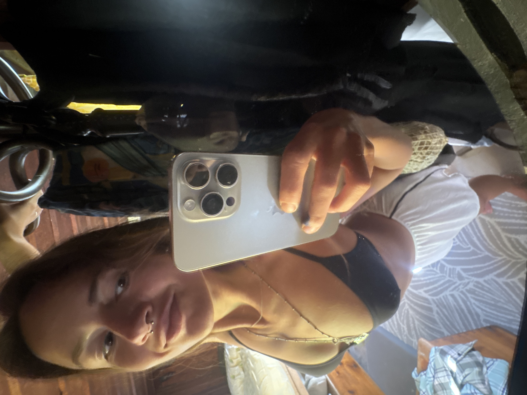 A young woman with a septum piercing and long brown hair is reflected in a mirror, taking a selfie with a smartphone. She is wearing a black bikini top and white shorts. Her right hand, holding the phone, is visible in the lower right portion of the frame. The background appears to be an indoor setting with wooden beams and fabric draped around what might be a closet or canopy. The lighting is warm and somewhat dim.