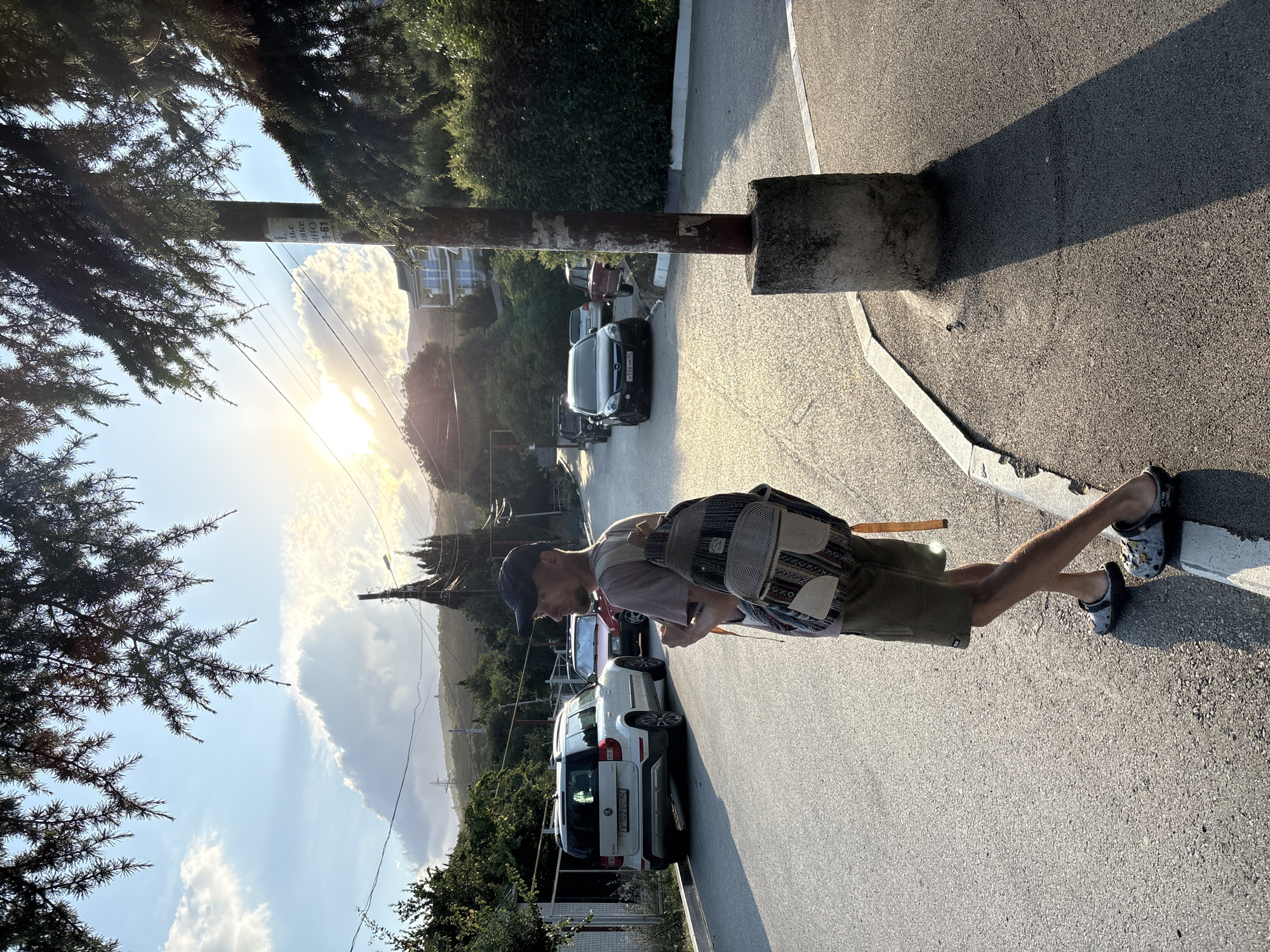 A man wearing a baseball cap, t-shirt, shorts, and Crocs walks on an asphalt road. He carries a patterned backpack with straps. The sun is setting, casting long shadows. Several cars are parked on the side of the road. In the background, there are trees and buildings on a hillside. A concrete pole with peeling paper labels stands on the right side of the road. The location is Malyi Mayak, Ukraine.