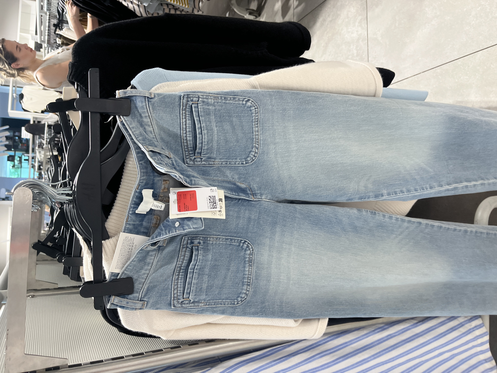 The image displays an indoor scene within a retail clothing store, set in Blagoevgrad, Bulgaria. In the foreground, a pair of light blue denim jeans hangs vertically from black plastic clip hangers. The jeans feature distinctive large, rectangular patch pockets on the front. Attached to the jeans are multiple labels, including a white tag, a red tag with partially obscured text, and a light brown tag. The brown tag clearly shows a QR code, various garment care symbols, and text in multiple languages, indicating manufacturing or care instructions.
Beneath the jeans, a stack of garments is visible, comprising a cream-colored ribbed sweater or top, a light blue top, and a black sweater or top. Further towards the bottom of the frame, a blue and white striped fabric is partially seen. In the upper left background, a woman with blonde hair, wearing a light-colored top, is partially visible as she appears to browse clothing on an adjacent rack. The setting includes metal clothing racks and bright, uniform interior lighting, indicative of a commercial retail environment. The activity captured is that of shopping or merchandise display within a clothing store.