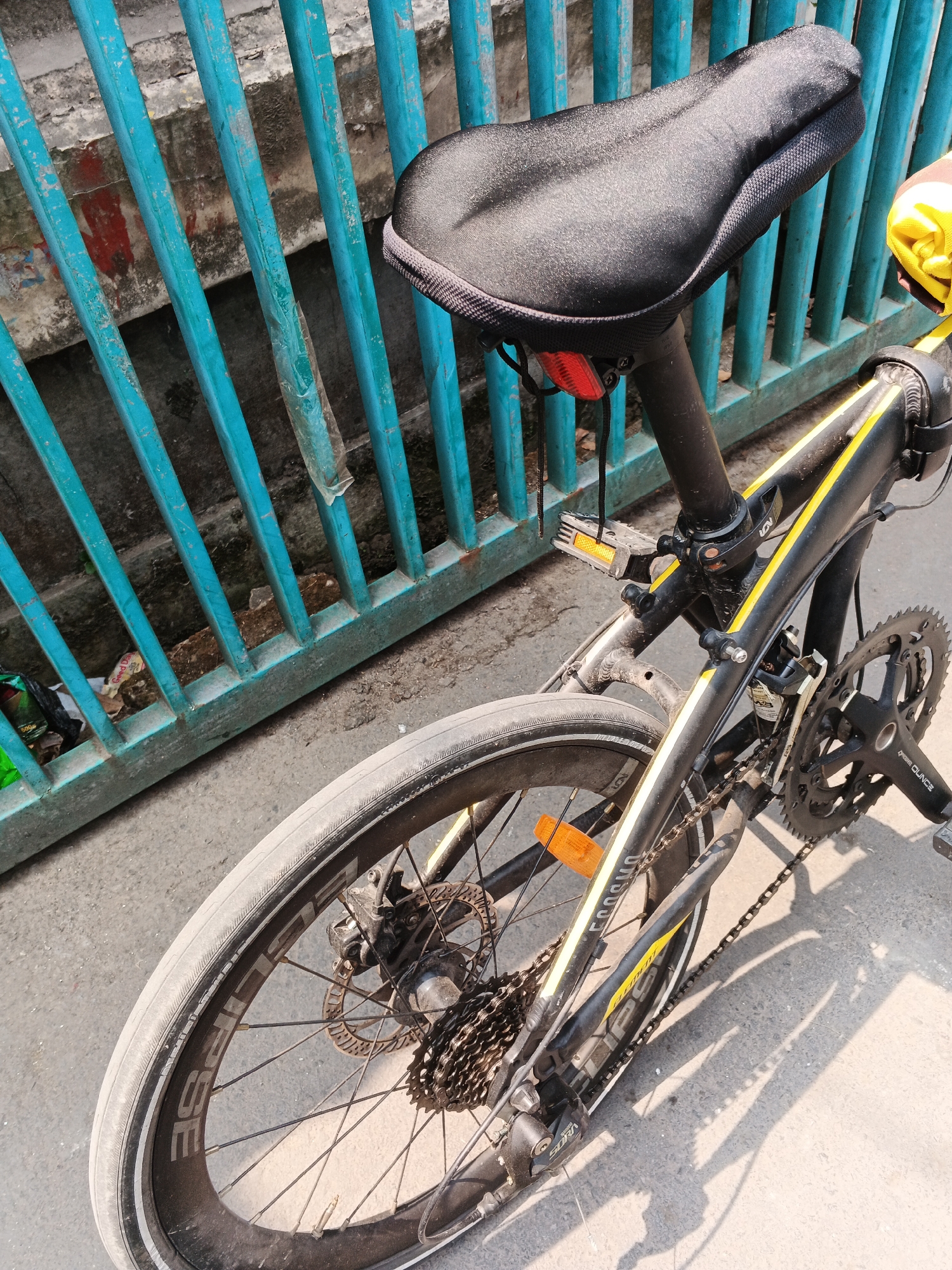 The image shows a close-up, high-angle view of the rear portion of a black folding bicycle parked next to a weathered, teal-colored metal fence. The bicycle has yellow accents on its frame and a black seat with a textured cover. Visible components include the rear wheel with a silver rim labeled "ECLIPSE," a cassette rear derailleur, a chain, and a crankset with black pedals. A red reflector is attached to the seat post, and an orange reflector is present on the frame near the rear wheel. The fence, made of vertical metal bars, appears to be old and slightly rusted in places, with some peeling paint. Behind the fence, a textured concrete wall with visible red and grey patches is present, along with some scattered debris on the ground. The scene is illuminated by sunlight, casting shadows on the asphalt ground. The location is identified as Gandasari, Indonesia.
