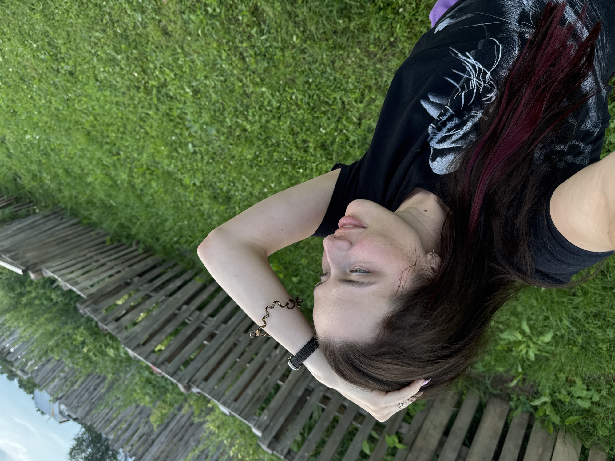 A medium shot shows a woman lying on her back in a grassy area next to a wooden fence. She is wearing a black t-shirt with a white graphic on it. Her left arm is bent at the elbow with her hand resting on her head, and she is wearing a black watch and a coiled bracelet on that arm. Her right arm is not visible. She has long, dark brown hair with streaks of burgundy. The woman is looking up and to her right. The background consists of a wooden fence, green grass, and some tall green plants behind the fence. The sky is visible at the top left of the image.
