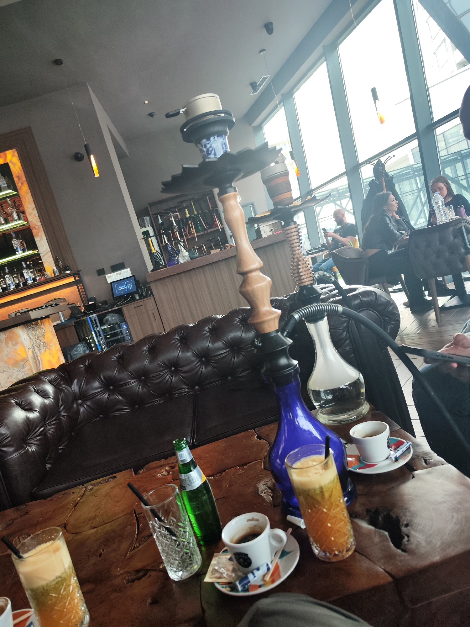 The image captures a scene in a cafe in Burgas, Bulgaria. A hookah pipe is prominently displayed on a wooden table, surrounded by several glasses and cups containing beverages. A green glass bottle is also visible on the table. A dark leather couch is positioned behind the table, extending towards the left side of the frame. In the background, patrons are seated at tables near a large window, with views of the cityscape. A bar area with shelves containing bottles is visible along the back wall. The lighting is a mix of natural light from the windows and artificial light from hanging fixtures.