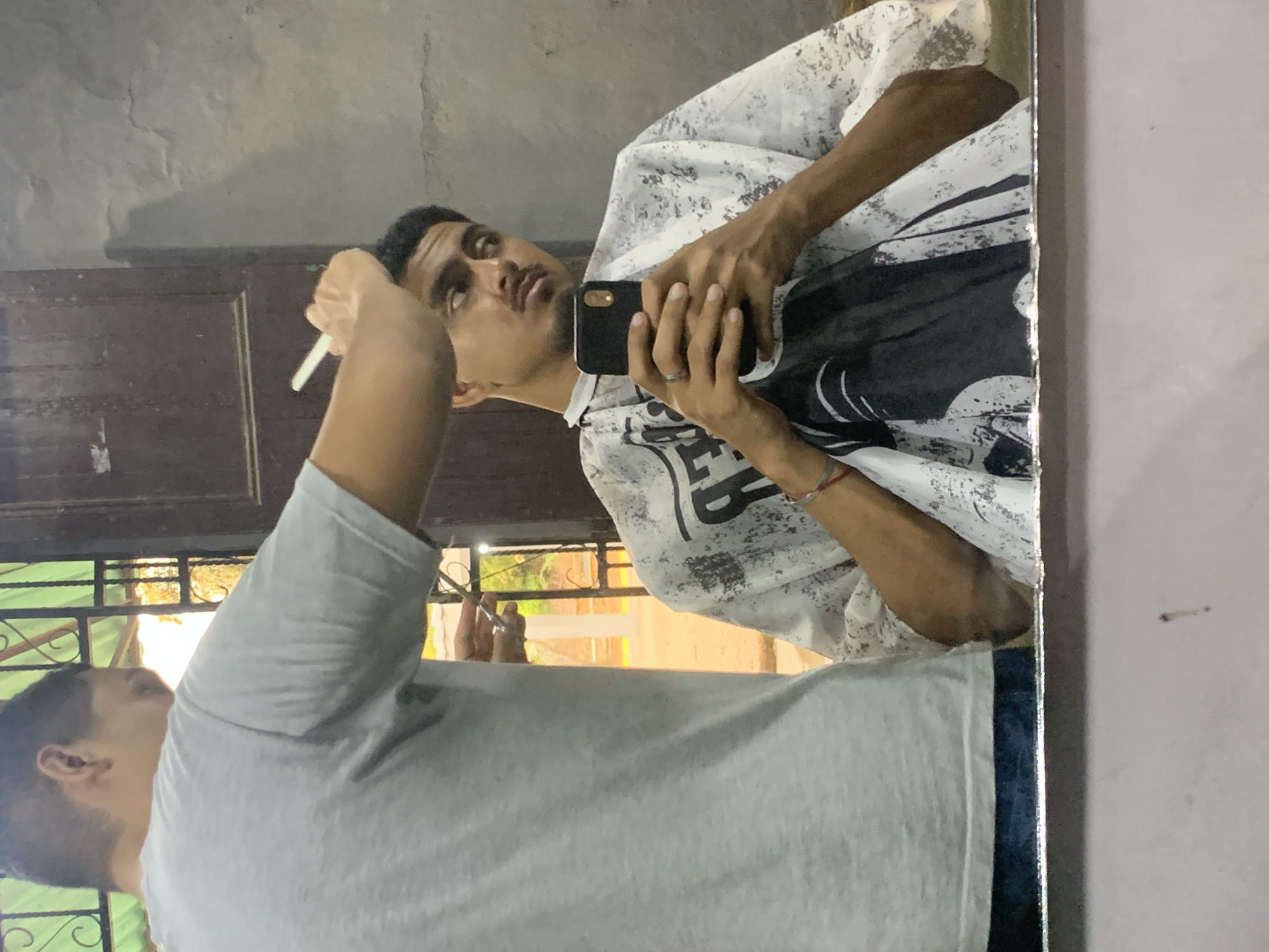 The image shows a reflection in a mirror, depicting two men in an indoor setting, likely in La Tagua, Venezuela. The central figure is a man seen in the mirror, wearing a white barber's cape with black abstract patterns, suggesting he is either getting a haircut or styling his hair. He is looking directly into the mirror with a neutral, focused expression. His left hand holds a black smartphone, which he is likely using to take the photo or for another purpose, while his right hand is raised, holding a slender white object that appears to be a comb or brush. He has short dark hair and a neatly trimmed beard. A ring is visible on his left ring finger.
Partially visible in the foreground, to the left, is the back and right arm of a second man, who is wearing a light grey t-shirt. His head is slightly turned, obscuring part of the reflected man's head. The background within the reflection shows a textured, light grey wall, a dark wooden door or cabinet, and through a window with dark metal grilles, some daylight and faint greenery are visible, indicating it is daytime. The overall scene conveys a casual moment, possibly a self-grooming session or a visit to a simple barbershop. No discernible text is visible on the patterned cape.