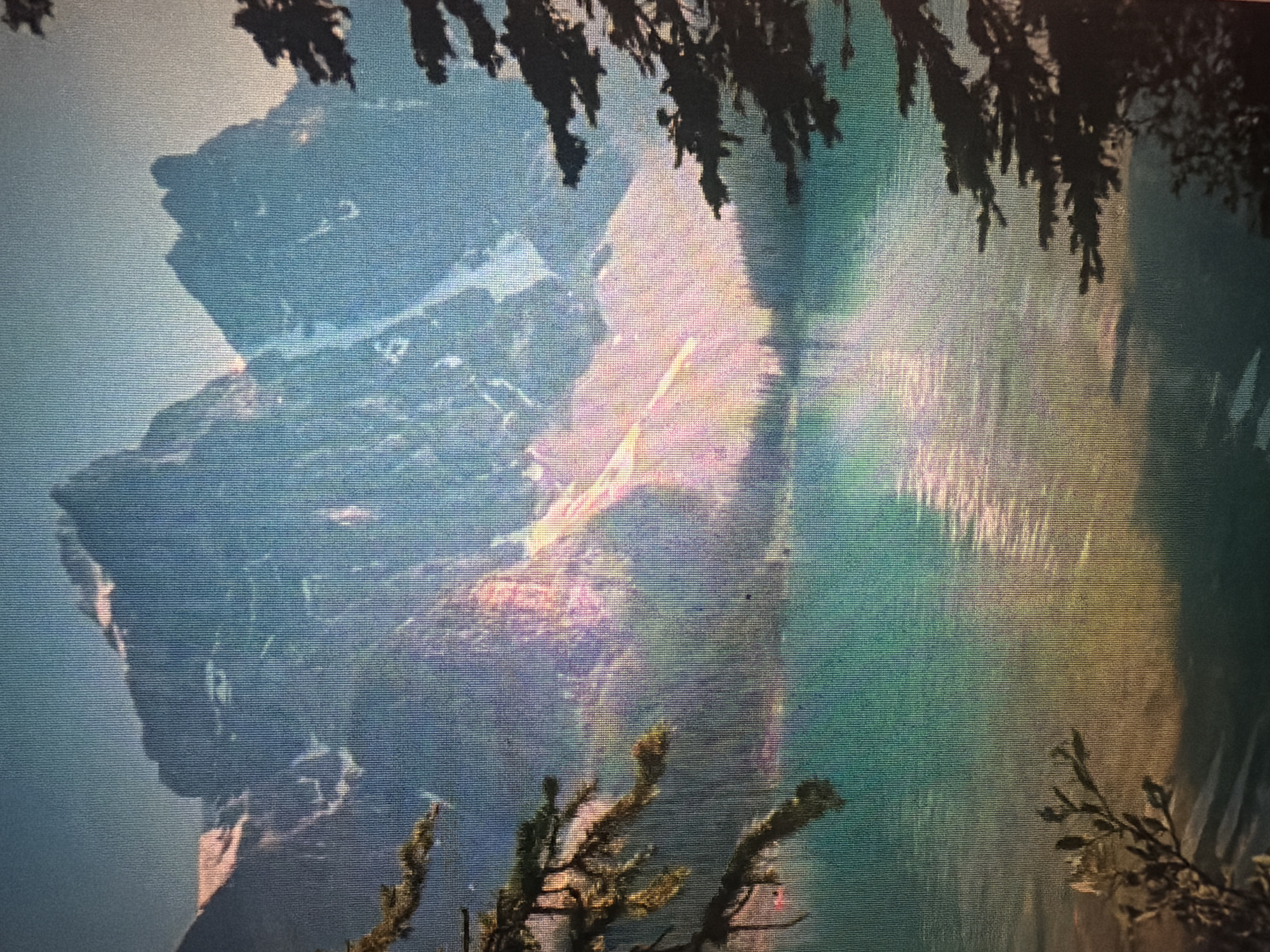 The image depicts a serene mountain landscape with a clear turquoise lake in the foreground. Majestic, snow-capped mountains rise in the background, bathed in the warm glow of sunlight, creating streaks of pink and orange on their rocky faces. Dark green pine trees frame the scene on both the left and right sides. The lake's surface reflects the light and the surrounding mountains, creating shimmering patterns. The overall atmosphere is peaceful and natural. This scene is not located in the city of Baan Mae Nam, Thailand.