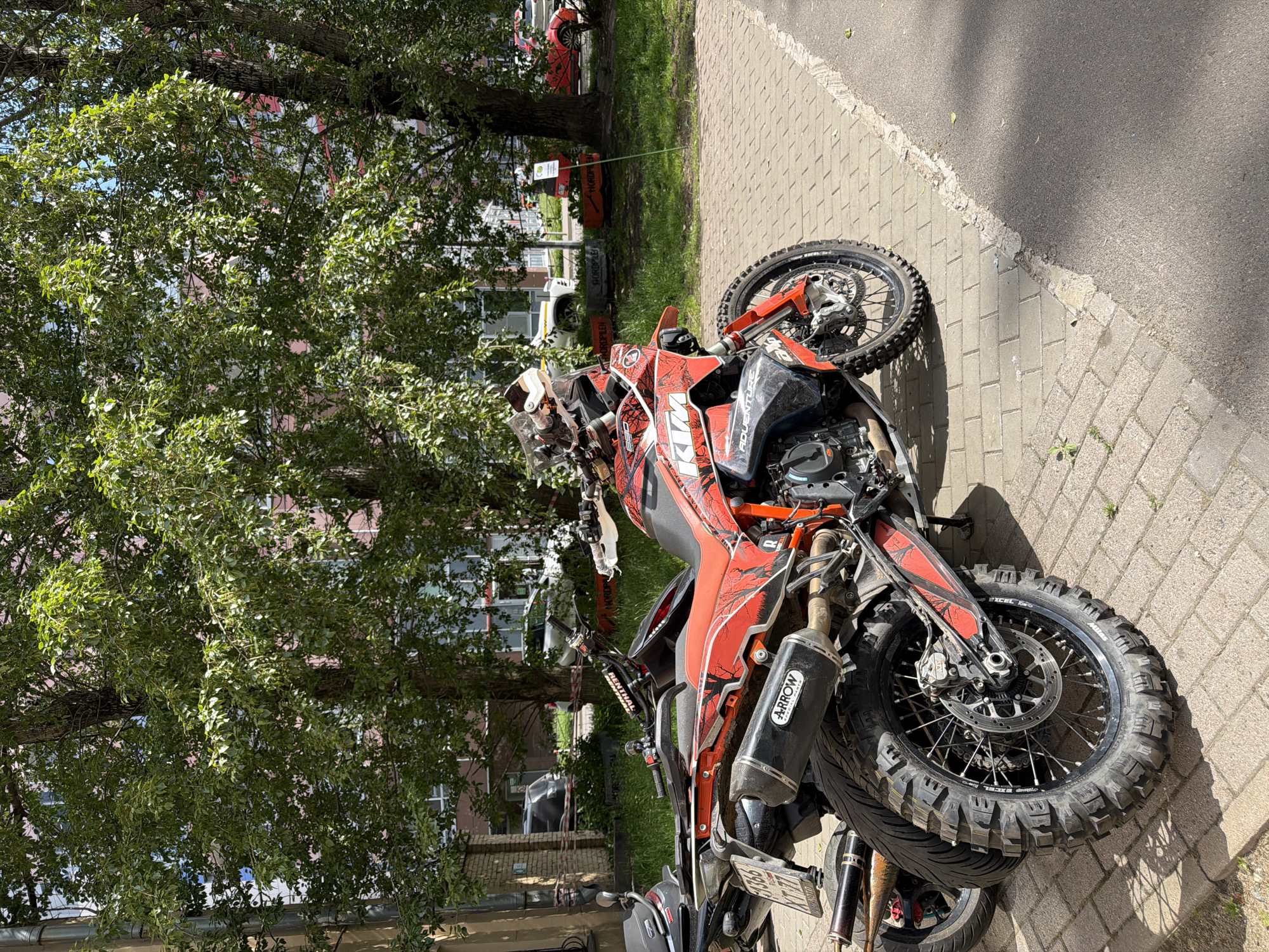 An orange and black KTM Adventure motorcycle is parked on a paved sidewalk in Saint Petersburg, Russia, on a bright, sunny day. The motorcycle is the central subject, featuring custom graphics, a prominent "KTM" logo on the tank, and "ADVENTURE" text on its side fairing. Notable details on the bike include a "Renthal" handlebar pad, an "Arrow" brand exhaust pipe, "Excel" rims, and "Mitas" tires with an aggressive tread pattern. The visible license plate, "X717BY 78", confirms the location in Russia.
The motorcycle is positioned on a sidewalk made of grey rectangular pavers. To its left, a large tree with dense green foliage occupies a grassy patch, partially obscuring the background. Beyond the tree and in the distance, residential buildings with multiple windows are visible. A red car and a white car are also partially seen further back, possibly on a street or parking lot. Several orange barriers, one of which appears to have "NORDPLEN" written on it, are situated near the tree. A thin green rope is also present in this area. The strong sunlight casts distinct shadows, indicating daytime. No people are visible, and no specific activity is taking place; the scene depicts a parked motorcycle in an urban or suburban setting.