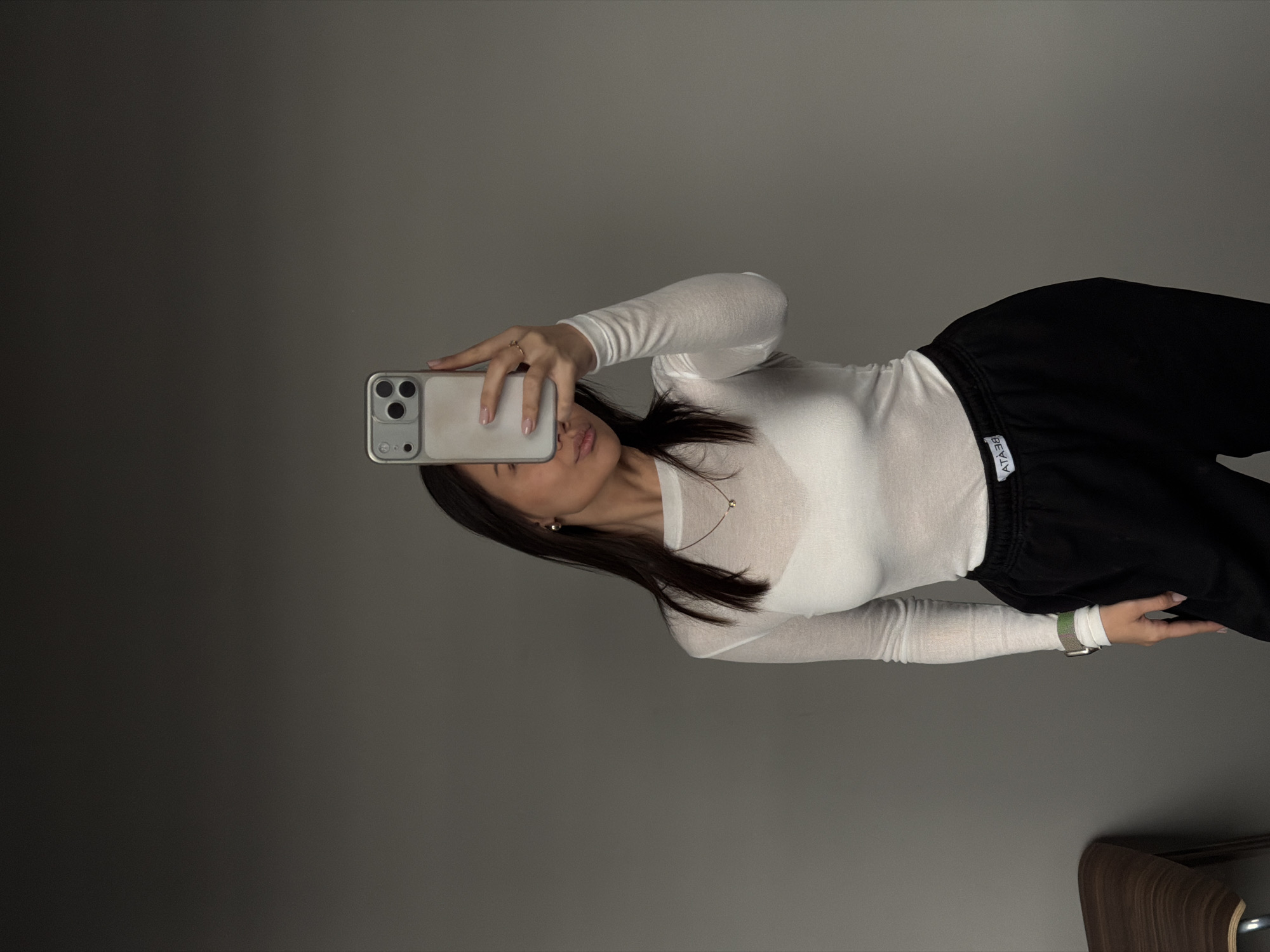 A young woman is taking a selfie with a smartphone. She is wearing a long-sleeved white top and black sweatpants. The background is a plain grey wall. There is a piece of wooden furniture visible in the lower right corner. The location is potentially Krasnoyarsk, Russia.