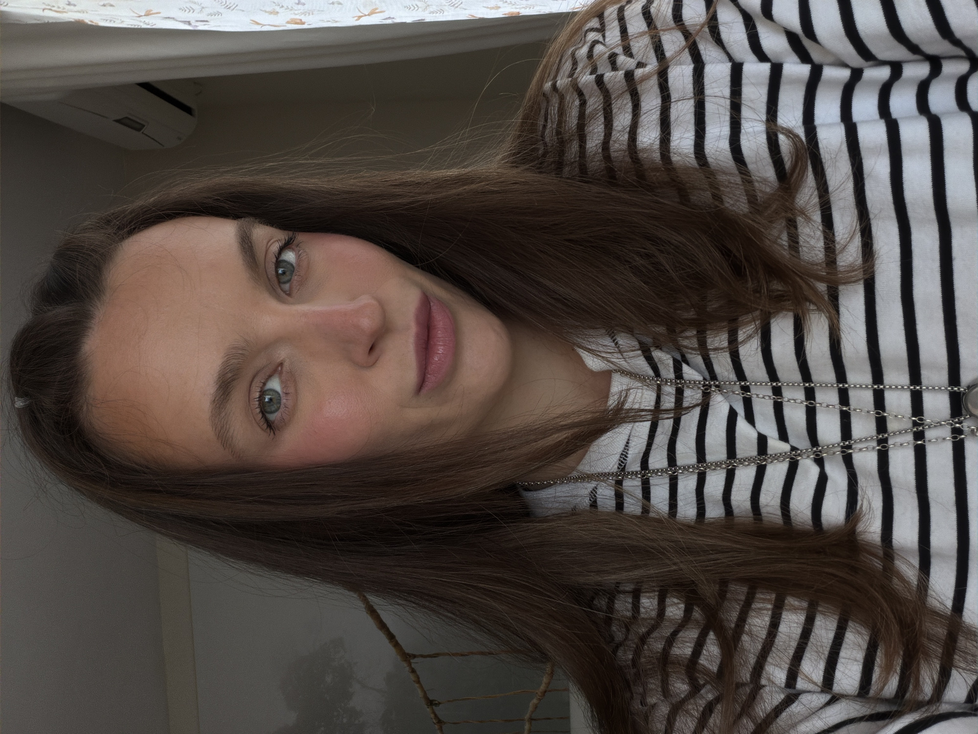 A woman with long, brown hair and fair skin is angled in the frame, facing the camera. She has light eyes and pink blush on her cheeks. She is wearing a black and white striped shirt, and a silver necklace is visible around her neck. The background shows a dimly lit room with a white wall and a patterned curtain above her head. A glimpse of the outside can be seen through a window, suggesting a neutral-toned landscape. The precise location in Budva, Montenegro, is not discernible from the visual details.