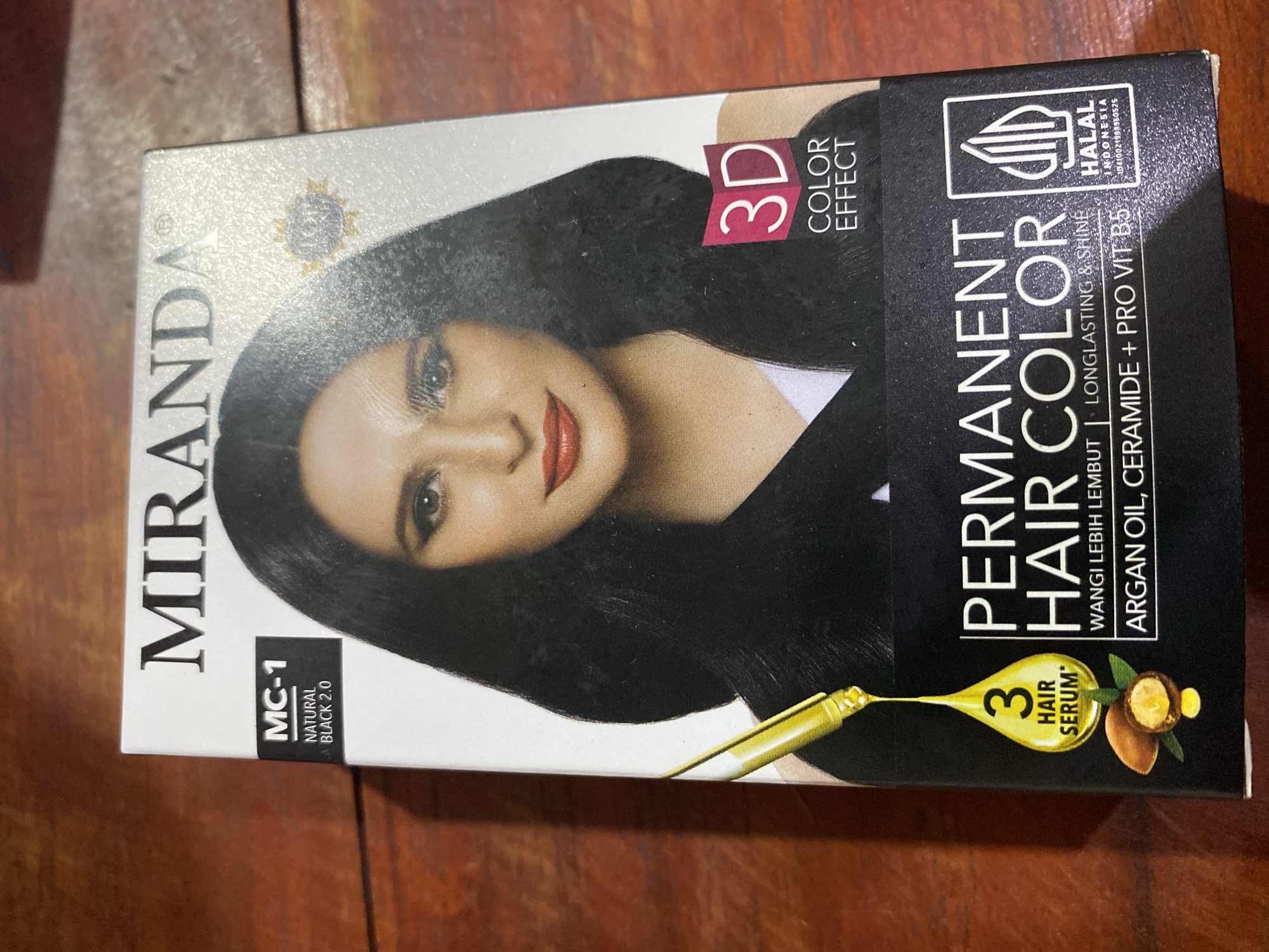 The image shows the front of a box of Miranda Permanent Hair Color, shade MC-1 Natural Black 2.0. The box features a portrait of a woman with long, dark hair and red lipstick. Text on the box indicates "3D Color Effect" and highlights "3 Hair Serum" with argan oil and pro-vitamin B5. A Halal certification for Indonesia is also visible. The box is placed on a wooden surface, likely a table, in what appears to be an indoor setting. There is no discernible information about its location within Ta', Indonesia.