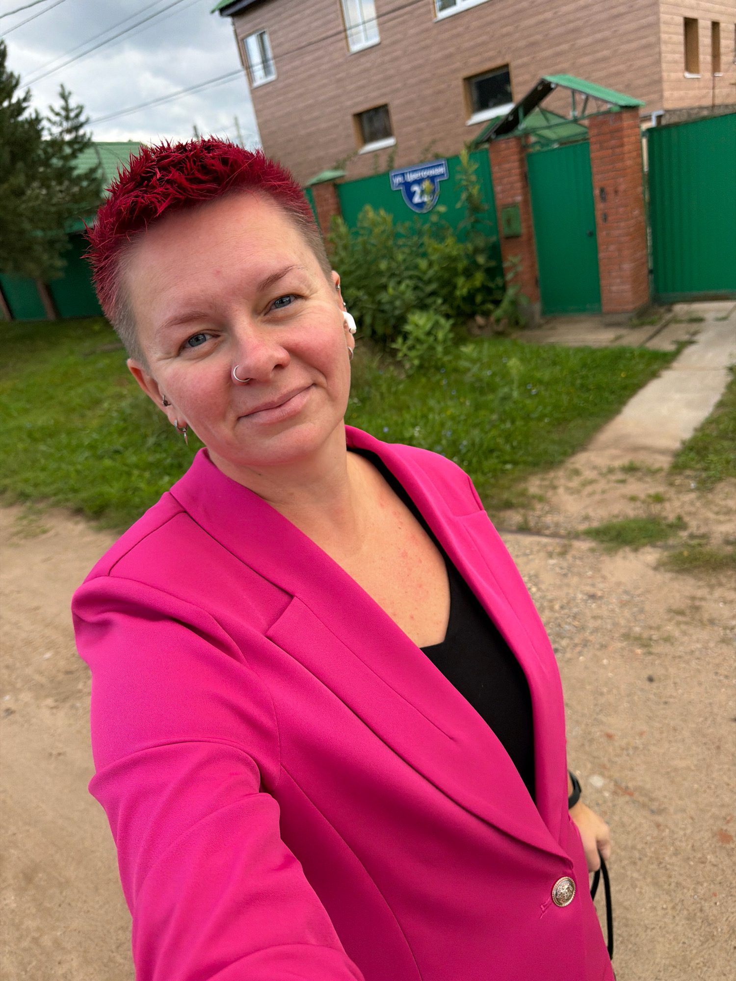 A woman with short, spiked, red-dyed hair and a nose ring smiles at the camera in front of a house with a green fence in Chaikovsky, Russia. She is wearing a bright pink blazer over a black top and white wireless earbuds. The house in the background has brown siding and a green roof. A blue street sign with the number "2" is visible on the fence. The ground is a mix of dirt and grass.