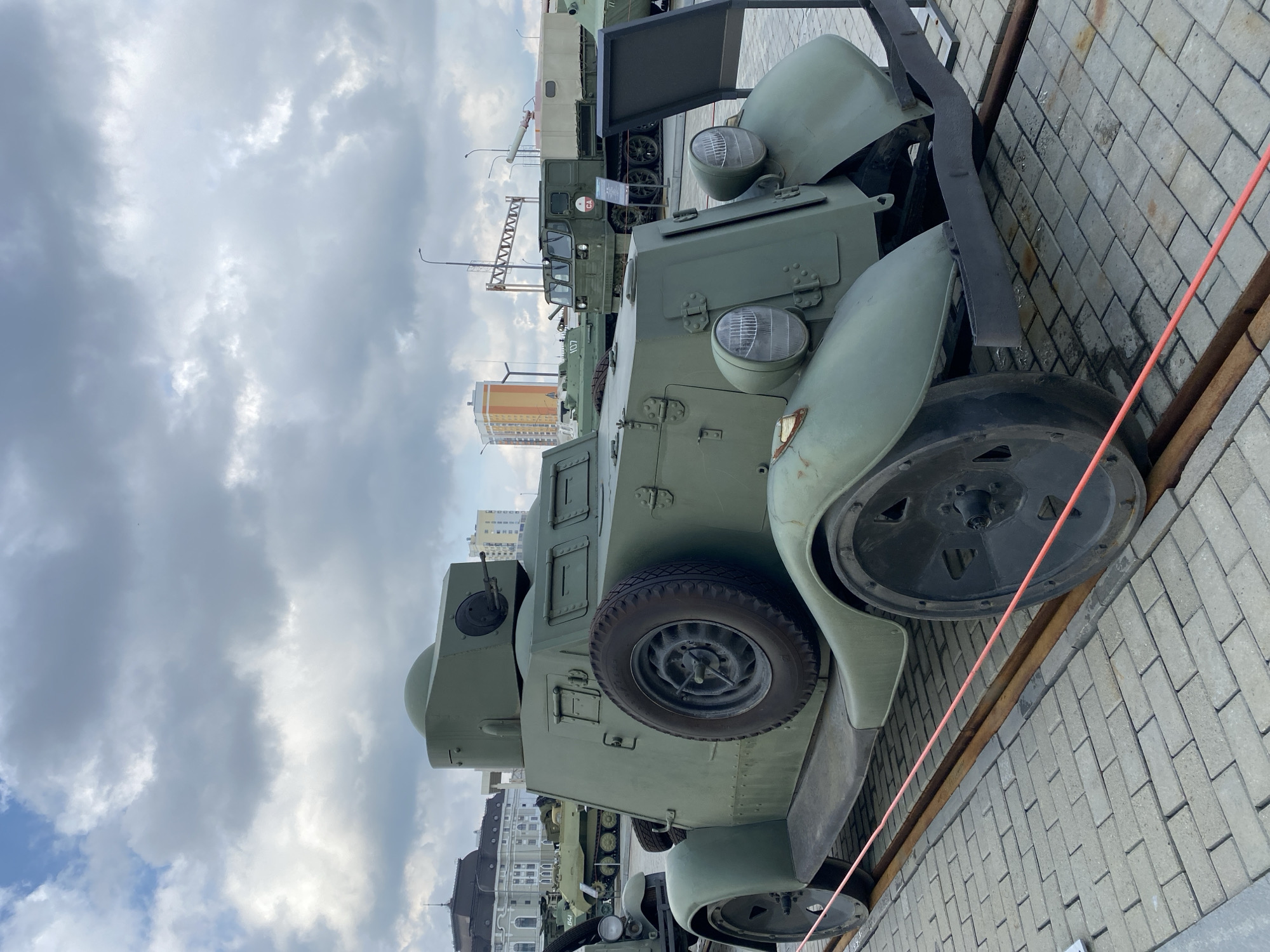 The image shows a military vehicle, likely a historical armored car, parked outdoors on a paved surface with what appear to be train tracks. The vehicle is painted a dull green and has a prominent spare tire mounted on its side, a turret with a visible machine gun, and large, distinctive headlights. In the background, other military vehicles, including a tank and a half-track vehicle, are visible, along with buildings and a cloudy sky. The scene is likely from the Military Technology Museum "Battle Glory of the Urals" in Verkhnyaya Pyshma, Russia, which features a collection of Soviet-era military hardware. There are no people visible in the image. The overall setting suggests an outdoor exhibition or museum display of historical military equipment.