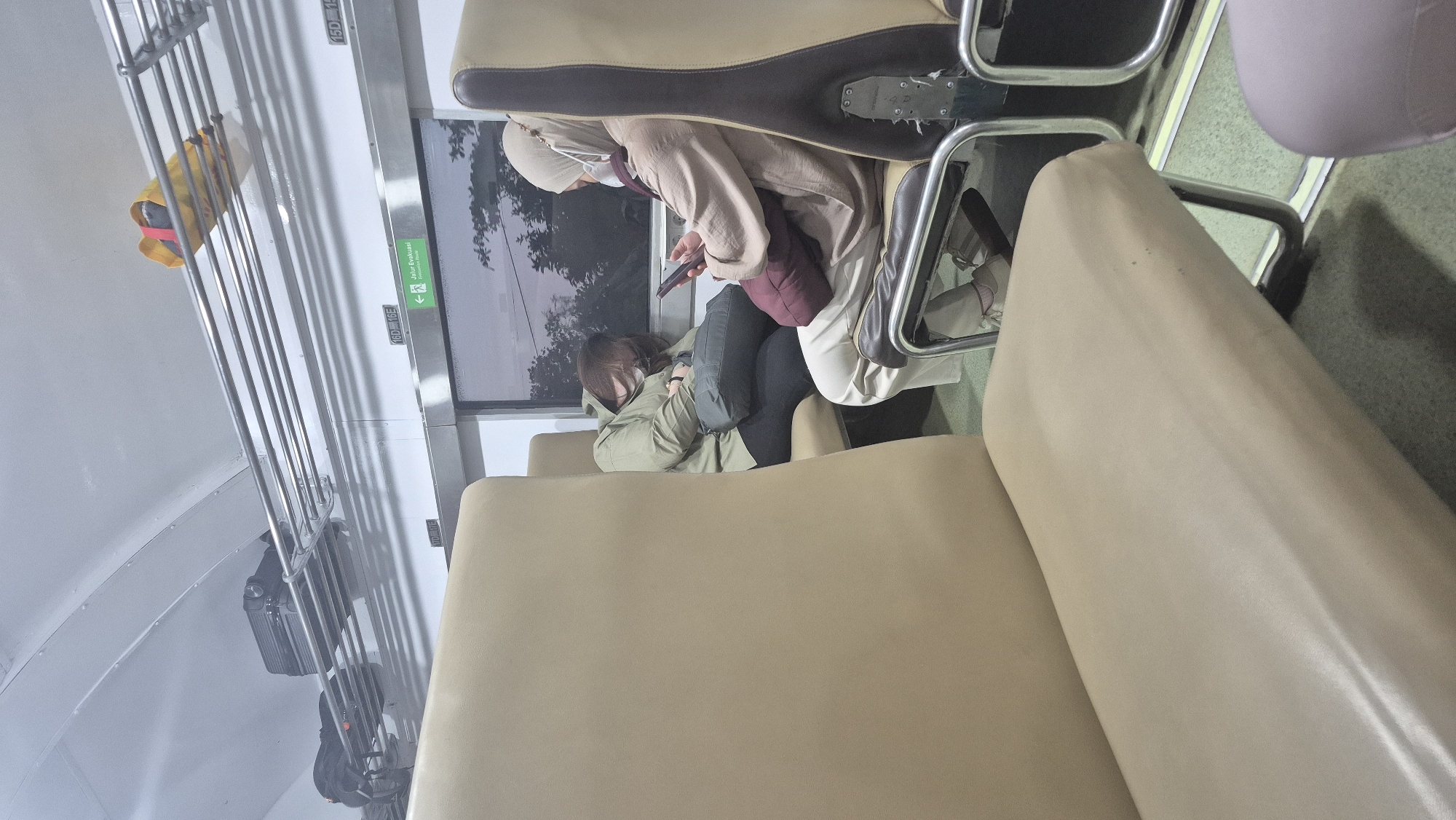 The image depicts the interior of a train carriage, likely in Sumuragung, Indonesia, based on the "Jalur Evakuasi" (Evacuation Route) signage. Two passengers are seated. In the foreground, the beige seat of an empty seat is visible. To the left, a passenger wearing a green jacket and black pants is asleep, leaning on a dark grey bag placed on their lap. They are also wearing a mask. To the right, a passenger wearing a beige hijab and a matching colored cardigan over white pants is looking at their phone. They are also wearing a mask and have a dark red pillow on their lap. Luggage, including a black suitcase and a backpack, is stored on overhead racks. A yellow bag is also visible on the rack. The window shows a dark, overcast sky with the silhouettes of trees. The overall lighting suggests it is either early morning or late evening.