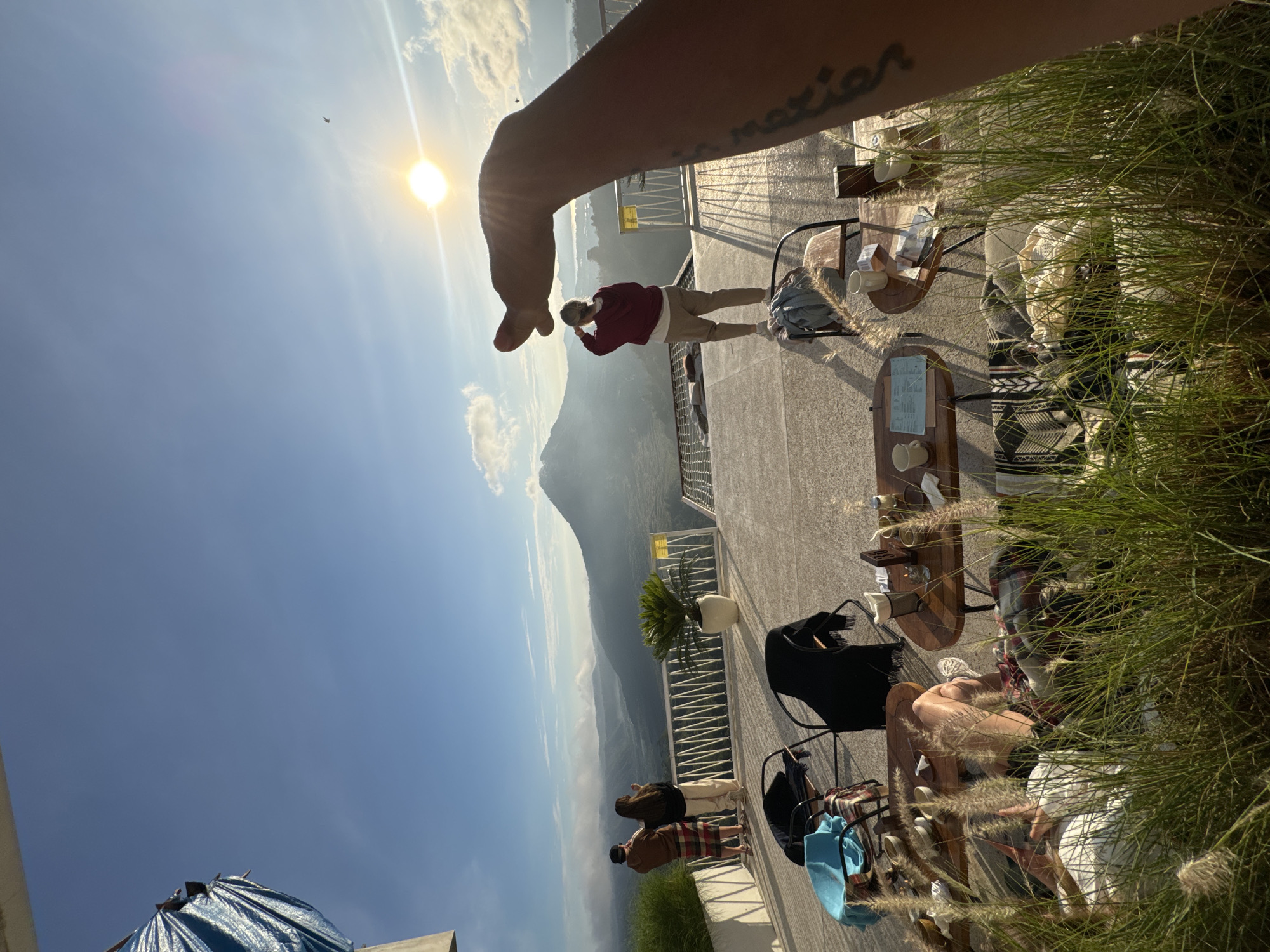 The image, seemingly taken on a sunny day in Kedisan, Indonesia, presents a panoramic view from an elevated platform, likely a restaurant or cafe. In the foreground, a person's arm with a tattoo is extended, partially obscuring the scene. The sun is high in the sky.
Below, a terrace or balcony setting is visible. People are present, seemingly enjoying the view. One individual, clad in a maroon shirt and beige pants, is taking a photo, presumably of the expansive mountainous landscape in the background. Tables and chairs are scattered around, suggesting a dining or relaxation area. Items like cups, plates, and menus adorn the tables.
In the far distance, the outline of a mountain can be seen, providing the location as being in the vicinity of the Kedisan region. Some grass is present.