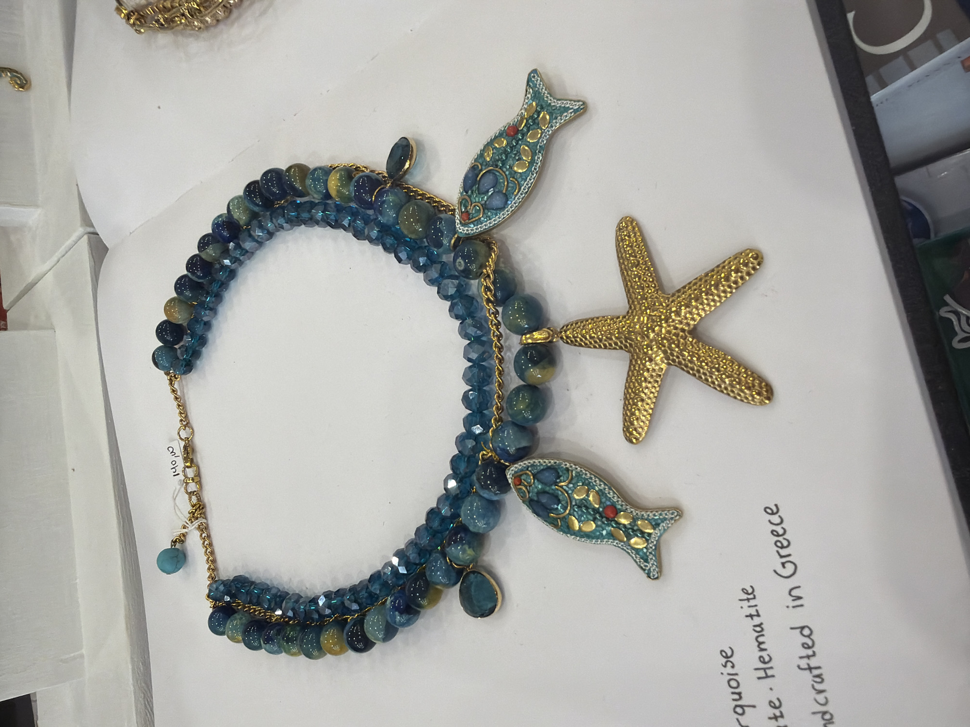 On a white surface, a beaded necklace with a gold starfish and two fish pendants is displayed. The necklace is made of teal, blue, and gold-colored beads, with gold chain accents. The fish pendants are blue with gold detailing and red accents. The starfish is gold. A handwritten label reads "Turquoise. Hematite. Handcrafted in Greece." The image was likely taken in Hydra, Greece, as indicated by the label.