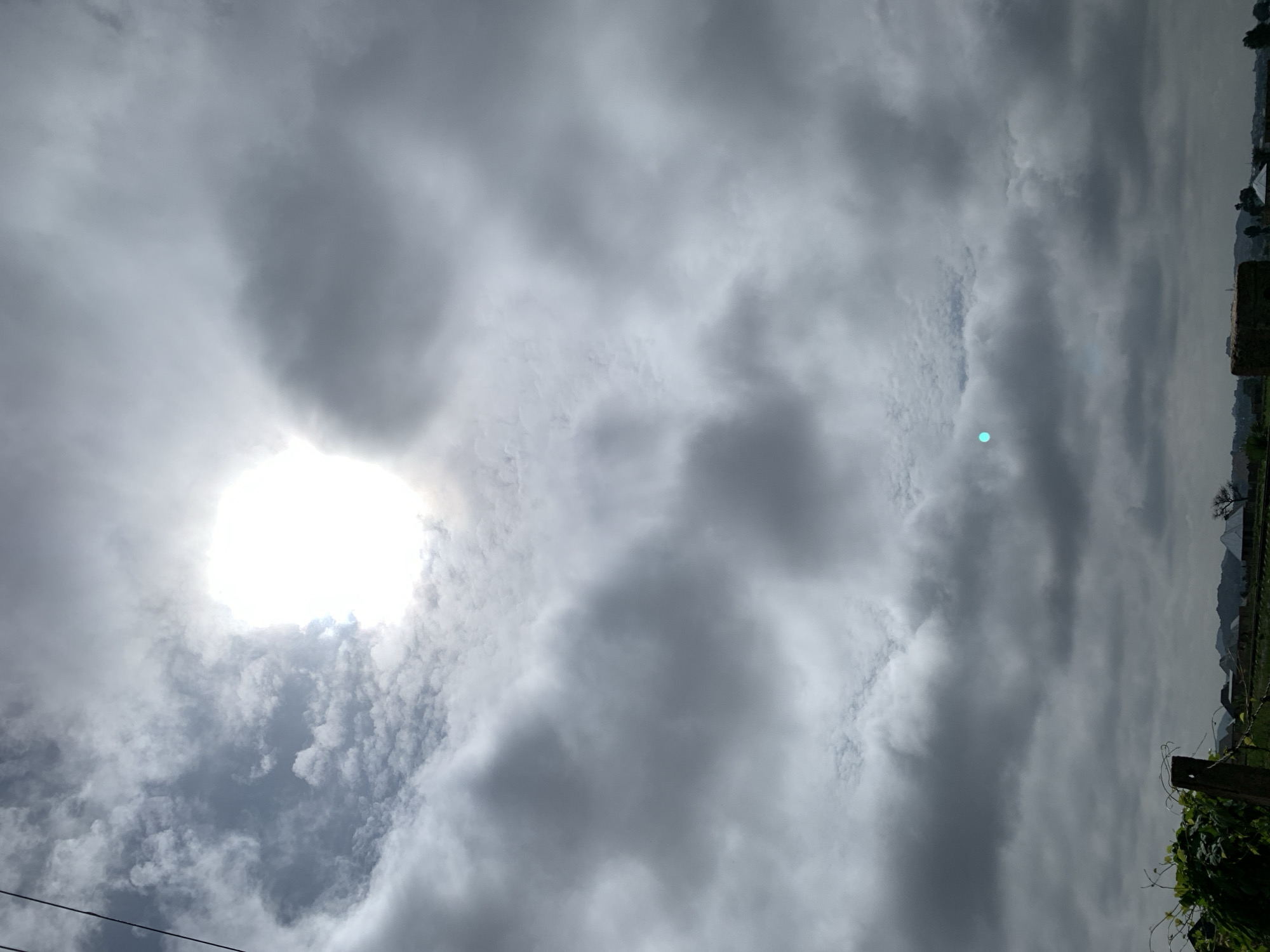 This is a low-angle, full-frame shot of a cloudy sky above Kuru Karama, Nigeria. The sun is visible, partially obscured by clouds, creating a bright, diffused light source in the upper center of the frame. A small, bright cyan circular artifact is present in the middle-left portion of the sky. The lower portion of the image shows a blurred landscape with vegetation in the foreground, including a wooden post with vines. In the distance, there are structures that appear to be houses and possibly a tree. The sky is dominated by large, dark, and light gray clouds, with some areas of thinner, white clouds closer to the sun. Electrical wires are visible in the upper left corner.