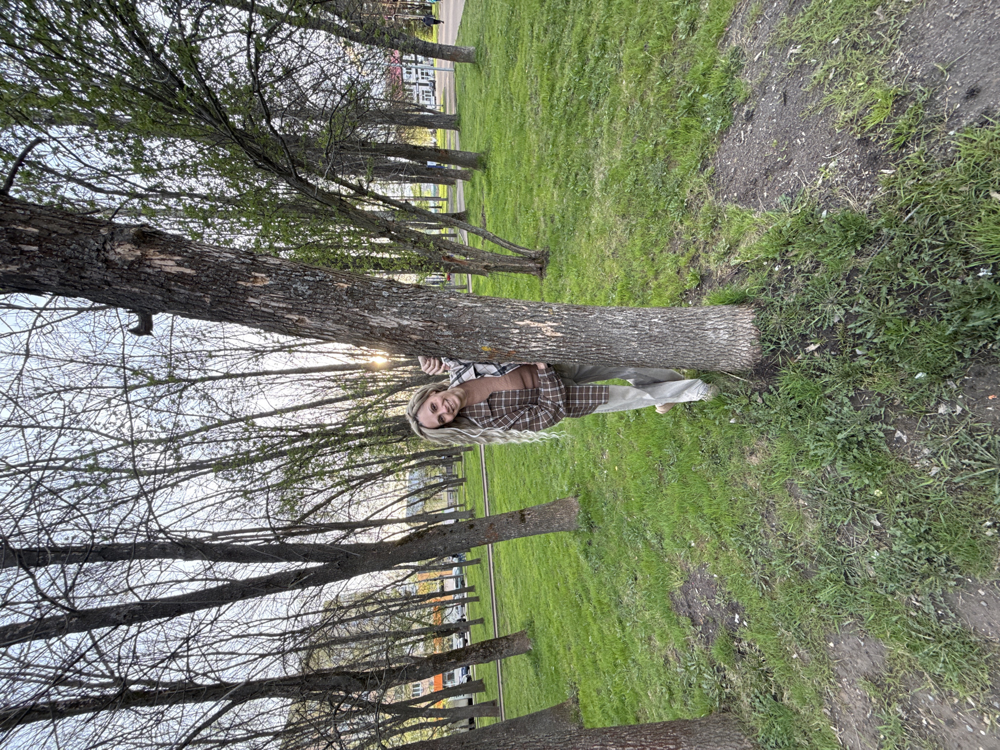 The image depicts a woman with long, blonde hair standing in a park in what is purported to be Rusanovo, Russia. She is positioned behind a tree, partially obscured by its trunk. The park is characterized by a row of trees lining the edge of a grassy area. In the background, buildings and parked cars are visible. The sky is overcast.