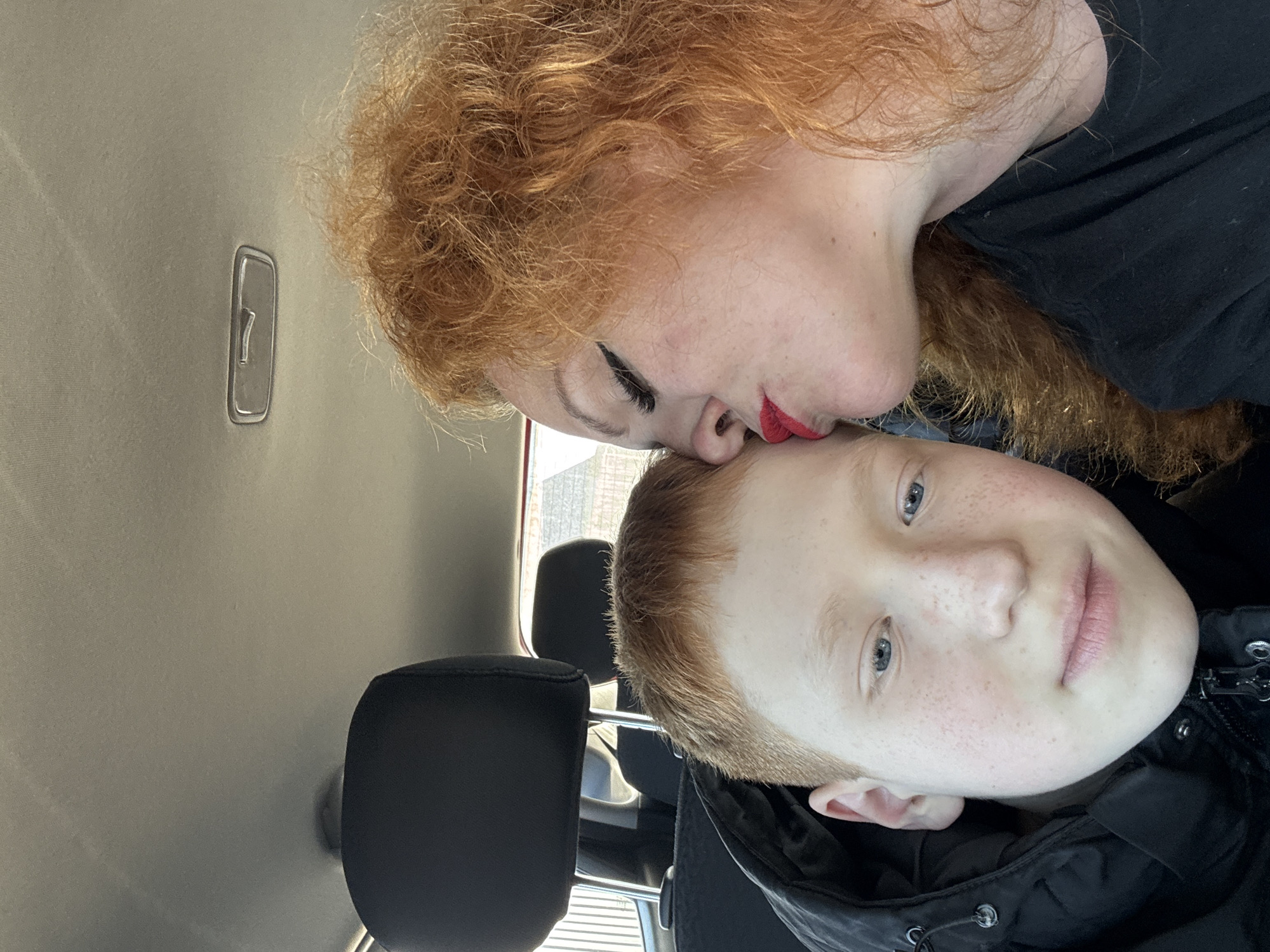 A woman with red curly hair and red lipstick is kissing a boy with red hair on the forehead. They are sitting in a car. The car has black headrests. A gray car ceiling is visible. The photo may be taken in Chaikovsky, Russia, but there is no verifiable information in the image to confirm that.