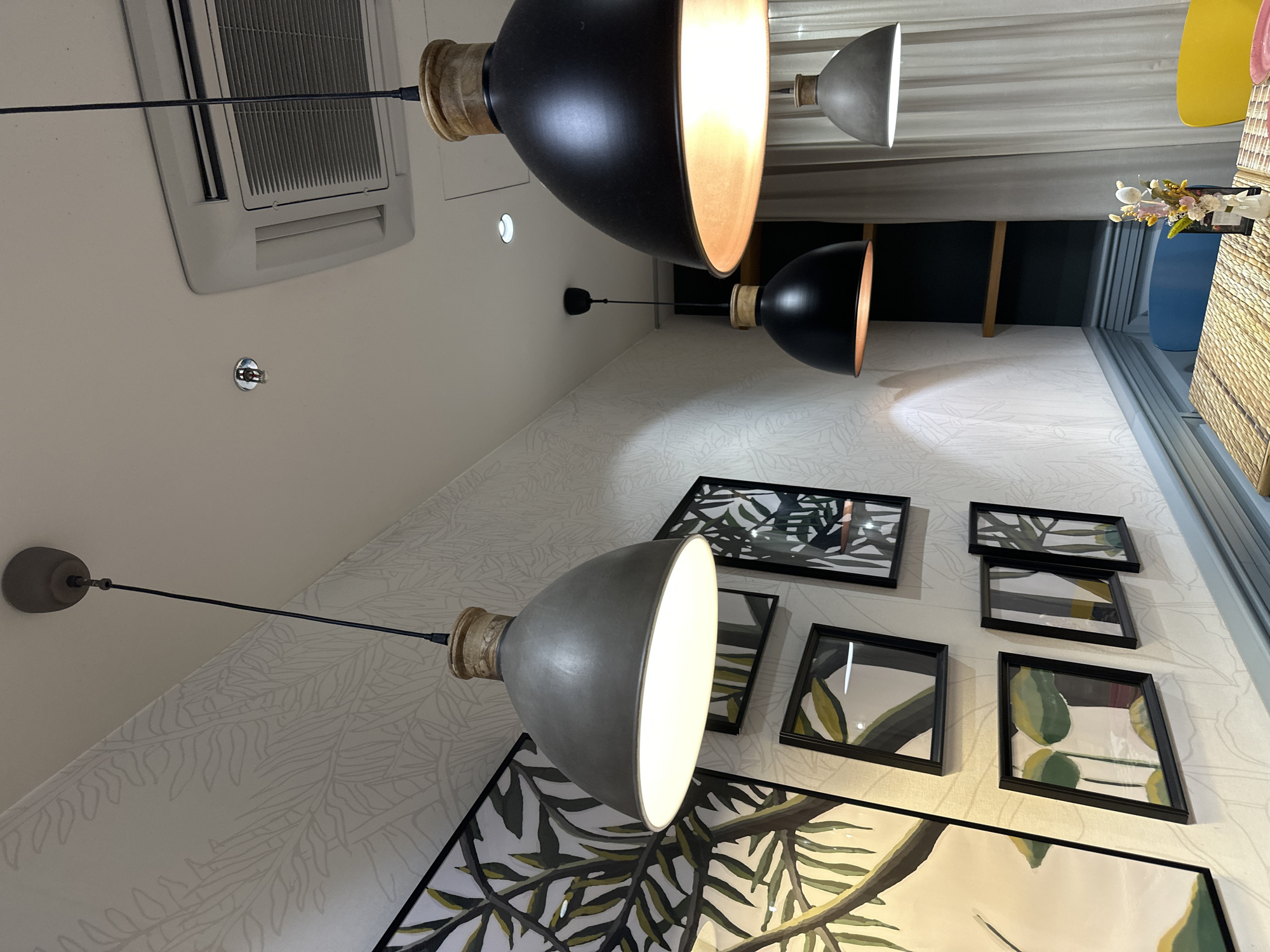 The scene is an interior space with a focus on lighting and wall decor. There are three pendant lamps, two with black shades and one with a gray shade. The lamps are positioned at different heights and distances from the camera. The wall is decorated with framed botanical prints. The background includes a glimpse of a window with curtains and possibly a glimpse of an outdoor space. This could possibly be located in Wrocław, Poland, given its specific mention, though the scene itself does not explicitly provide locational data. The specific details of the location within the city and any people present are not visible in the image.