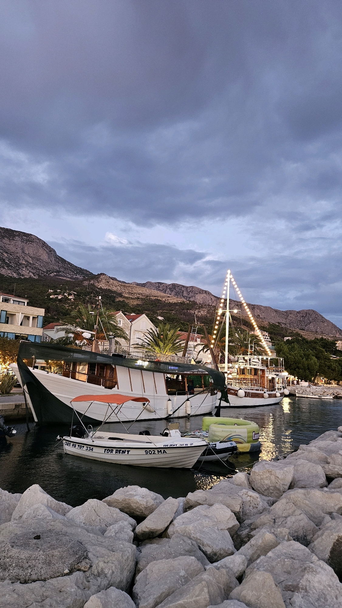 The image depicts a serene harbor scene in Općina Tučepi, Croatia, at dusk. A large, white boat with an orange canopy is docked, alongside a more ornate, larger boat adorned with a string of lights that trace its mast. In the foreground, a cluster of large, grey rocks leads down to the water's edge. The water is calm, reflecting the twilight sky and the lights of the boats. Behind the boats, a hillside town with white buildings and red roofs is visible, nestled against a backdrop of dark, imposing mountains. Palm trees are scattered throughout the landscape, adding a tropical feel to the Mediterranean setting. The sky is overcast with shades of grey and purple, indicating the transition from day to night. The overall atmosphere is tranquil and picturesque.