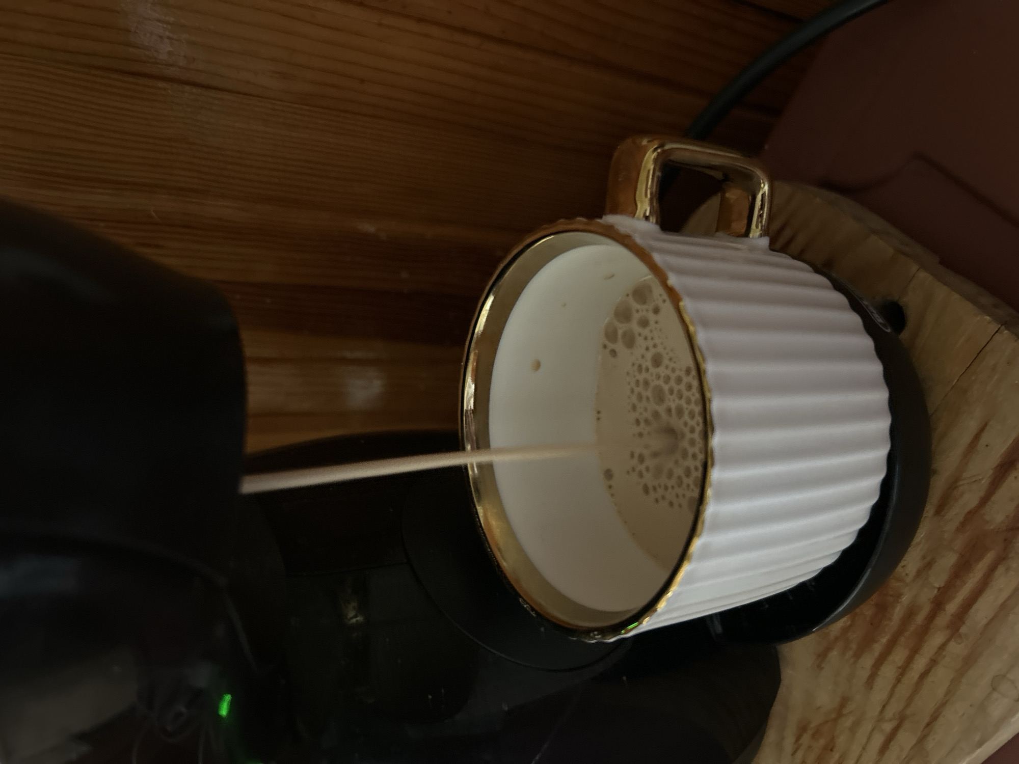 In Basarabeasca, Moldova, a coffee machine dispenses a light brown beverage into a white, ribbed mug with a gold rim and handle. The mug sits on a dark saucer atop a wooden surface. The background features a dark appliance and a wooden wall with vertical planks. Green indicator lights are visible on the appliance.