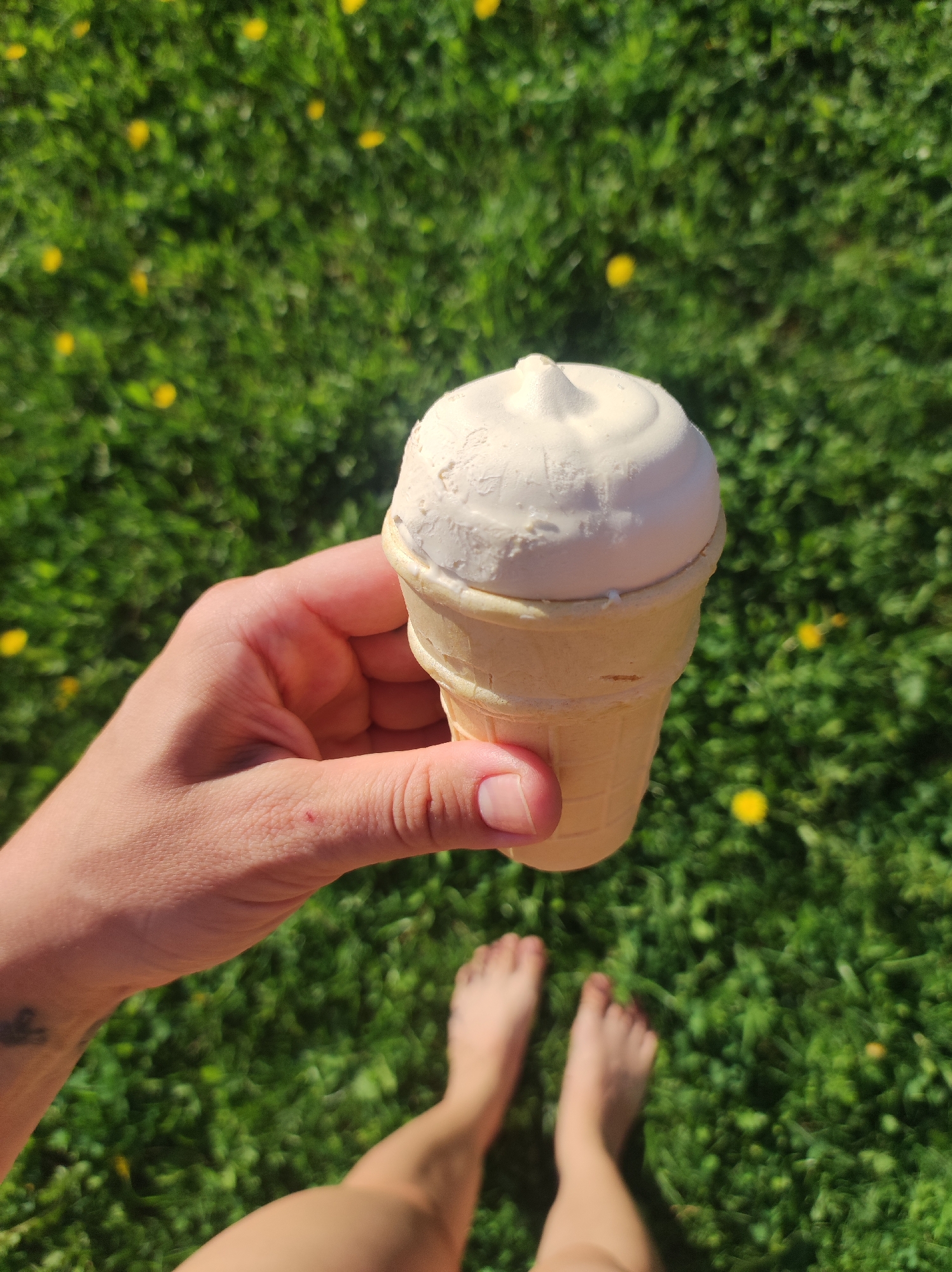 A person is holding an ice cream cone in their hand, standing barefoot on a grassy lawn. The ice cream is white and swirled on top of a light tan cone. The grass is green with small yellow flowers scattered throughout. The person's hand is holding the ice cream cone, and their feet are visible in the lower portion of the image. The location cannot be verified as being in Фоки, Russia.