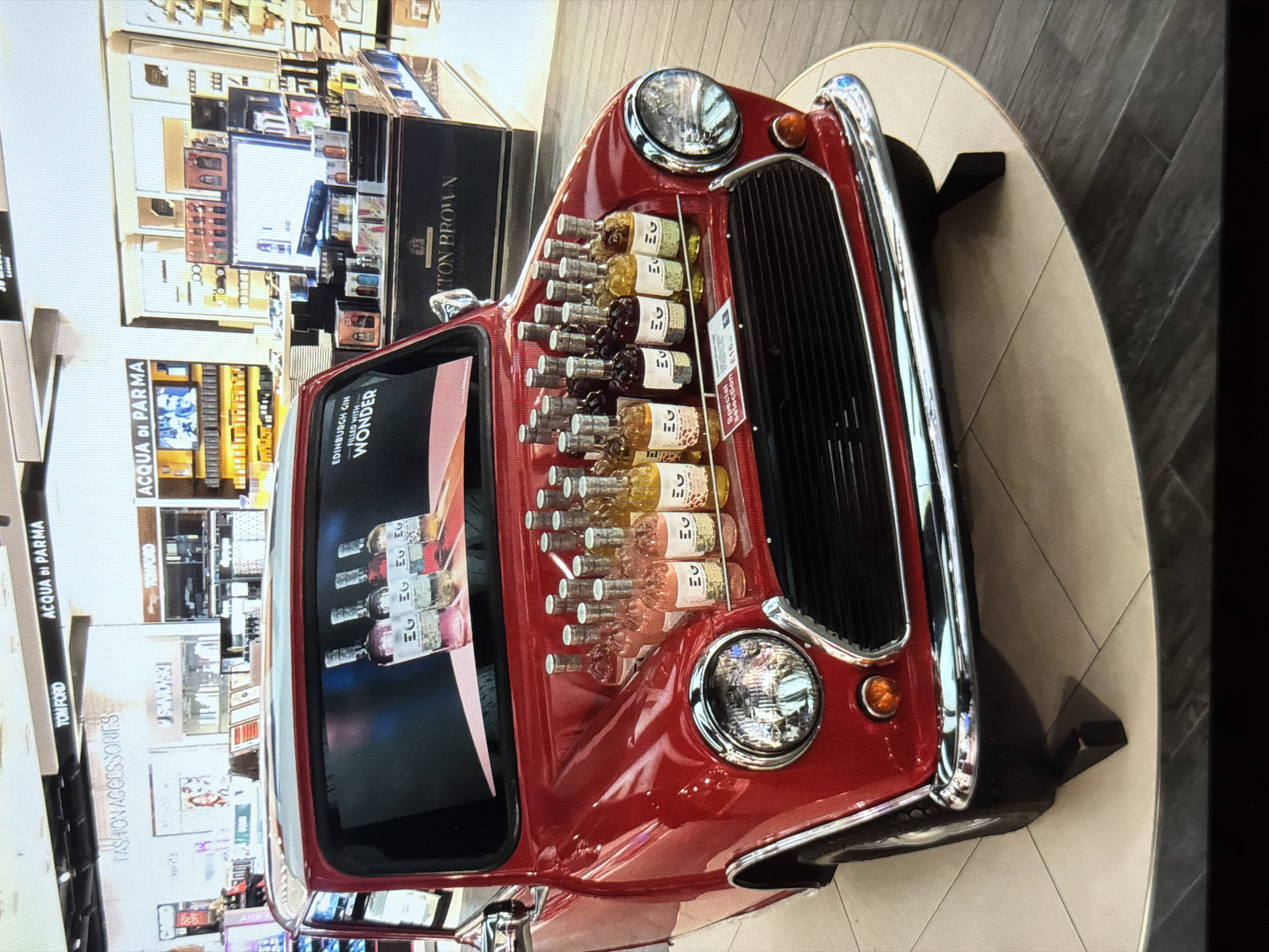 This indoor shot features a red classic Mini Cooper car, repurposed as a display stand for Edinburgh Gin, a brand promoted on the car's windshield. The vehicle is parked on a circular, light-colored platform. The car's hood is adorned with rows of gin bottles, each labelled "EG". The display includes a sign advertising a "Special selection" for £16. The backdrop consists of store displays featuring brands such as "ACQUA DI PARMA", "TOM FORD", and "LTON BROWN", suggesting a luxury retail environment. The exact location within the city of Burgas, Bulgaria, cannot be determined from the visual information provided.