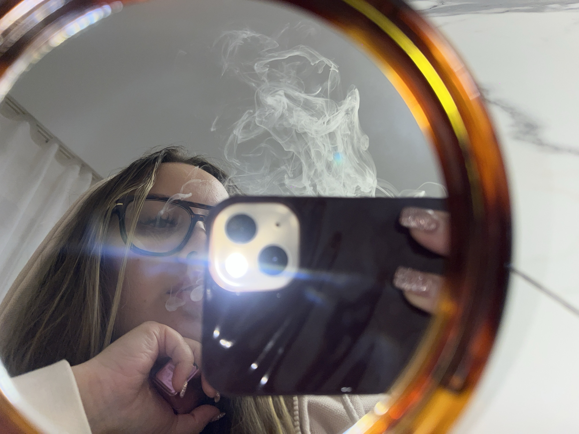 The image displays a mirror selfie of a person, likely a woman, taken indoors. The shot is captured through a round mirror featuring a distinct tortoise-shell frame. The person is holding a smartphone, identifiable as an iPhone due to its camera arrangement, in their right hand, with its bright flash illuminating the scene and creating prominent lens flares.
The individual has long, light brown hair and wears dark-rimmed glasses. Their face is partially obscured by the phone and the angle, but a significant plume of white smoke or vapor is seen emanating from their mouth, indicating they are either vaping or smoking. In their left hand, they hold a small, pink or purple electronic device, possibly a vape pen or a small lighter. Their visible fingers feature glittery nail polish.
The background reflected in the mirror consists of light-colored walls; the right side shows a subtle marble pattern, while the left side reveals a white curtain or soft furnishing. The indoor setting offers no specific time of day or weather cues. Although given the location context of Krasnodar, Russia, the visual details within the image itself are generic and do not provide specific geographical indicators. No discernible text is visible on any objects within the frame.