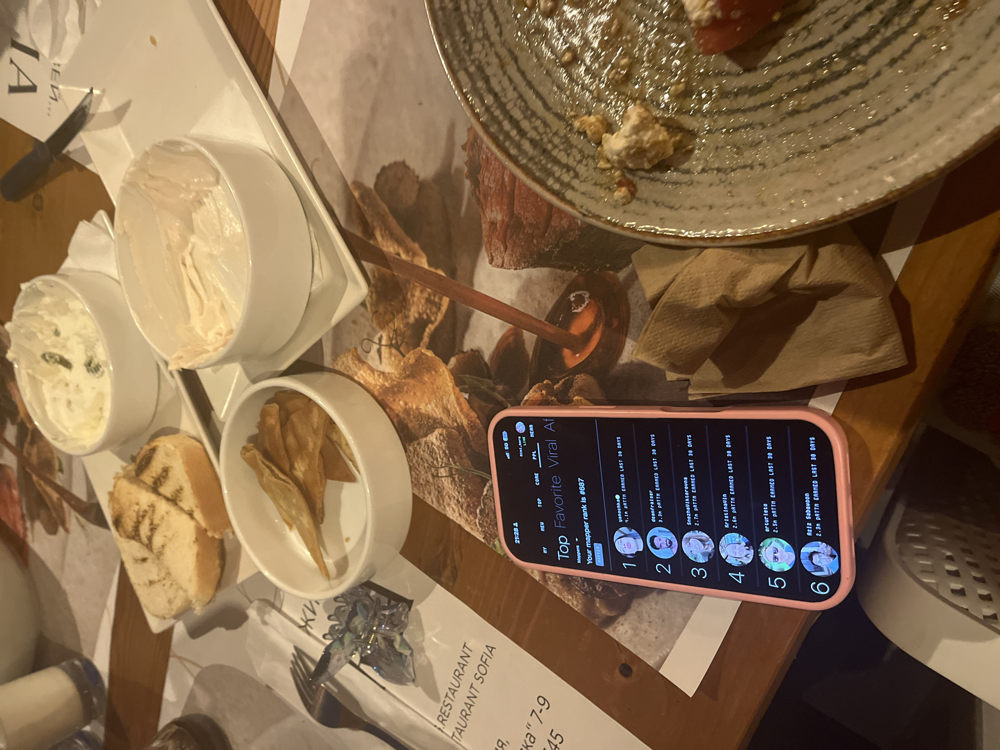 The image depicts a close-up, eye-level view of a table setting at a restaurant, likely in Tsarevo, Bulgaria, given the presence of a menu with Cyrillic script referencing "RESTAURANT SOFIA" and "Tsarevo." The scene is dominated by various food items and a smartphone displaying a ranking list.
In the foreground, a smartphone with a pink case is placed on a textured surface, possibly a wooden table, displaying a list titled "Top Favorite Viral Af" with user rankings and earned points. The screen shows "Your mapper rank is #687."
To the left of the phone, a white ceramic bowl contains what appears to be lightly toasted bread slices. Next to this bowl is another white ceramic bowl filled with thin, crispy fried snacks, possibly chips. Further to the left, a fork with a decorative, crystalline object is visible.
In the background, a white ceramic bowl contains a creamy, white dip with green specks, possibly a herb dip or spread. Adjacent to it is another white ceramic bowl containing a small amount of a pinkish-white substance, suggesting it has been partially consumed.
To the right of the smartphone, a partially eaten plate with food remnants is visible. The plate has a patterned rim and contains crumbs, a red item, and white crumbled pieces, possibly cheese. A folded brown napkin is placed next to the plate.
The overall environment appears to be an indoor dining setting, with warm lighting and a focus on the presented food and the smartphone's content. The image captures a moment during a meal, with evidence of consumption on the plate and in the bowls. The backdrop features a patterned tablecloth or placemat with imagery of food.
