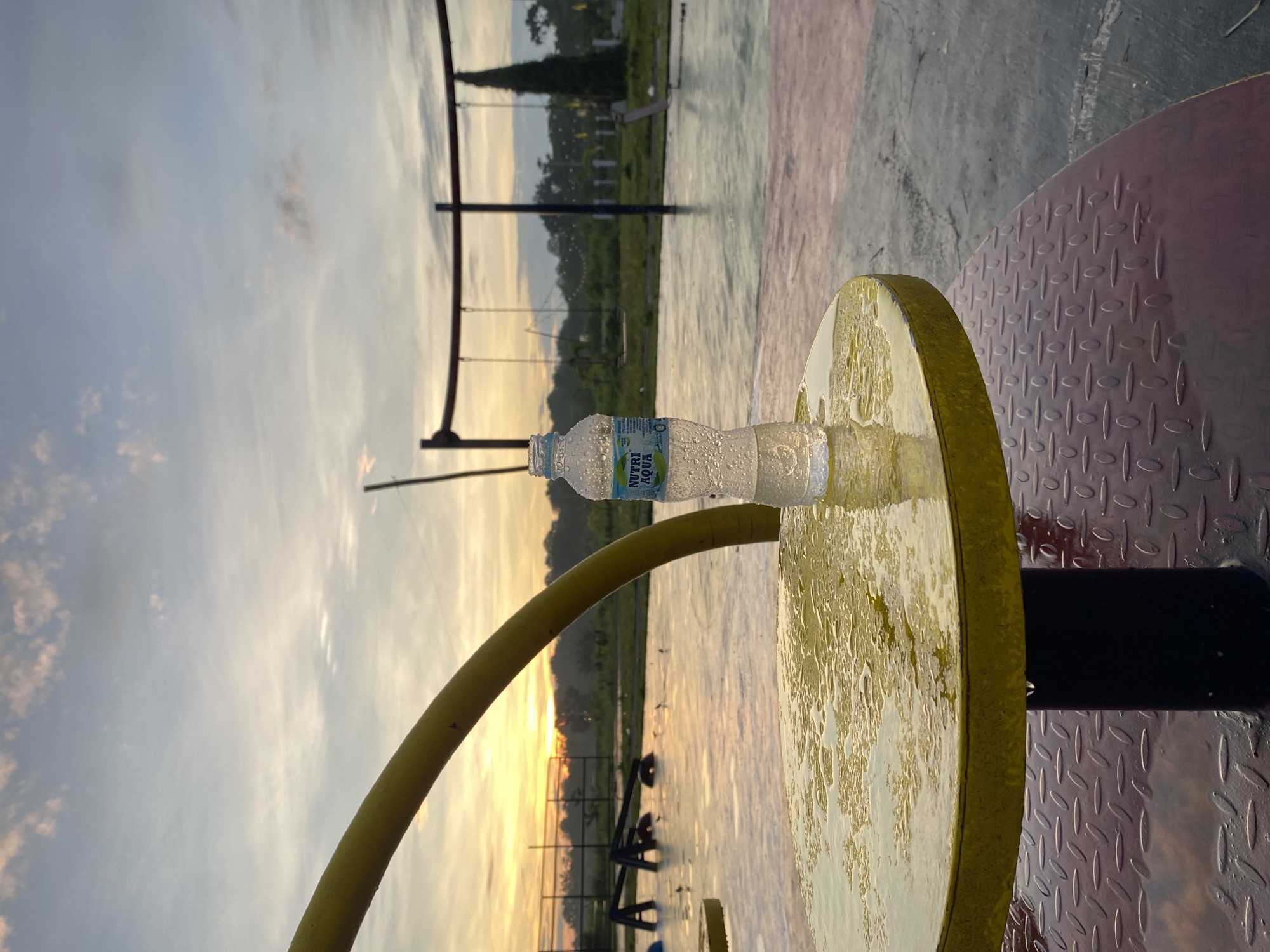 The image displays an outdoor scene captured during either sunrise or sunset, evoking a tranquil and serene atmosphere. In the foreground, a clear plastic water bottle, heavily covered in condensation, lies horizontally on a damp, circular, yellow-painted metal disc. Adjacent to this, on the right, is a dark red or purple diamond-plate metal surface, also appearing wet and reflective. A thick, curved yellow pipe or hose diagonally traverses the frame, extending from the lower-left towards the upper-right.
The background reveals a spacious outdoor area, possibly a park, playground, or a sports court in Davao City, Philippines. The ground, which appears to be a paved or hard surface, is noticeably wet and reflective, mirroring the soft, golden light from the low sun on the horizon. Silhouetted against the sky are various structures, including what seem to be playground swings, fence-like elements, and a distinctive tall, dark, conical evergreen tree positioned on the left side of the background. The sky transitions from a soft blue overhead to a warm, glowing yellow and orange near the horizon, typical of dawn or dusk.
No individuals are visible, and no specific activity is taking place; the scene is one of stillness. The heavy condensation on the water bottle and the wet surfaces of the ground and foreground objects strongly suggest humid conditions, recent rain, or perhaps heavy morning dew.
The water bottle's label clearly displays the brand name "NUTRI AQUA," with additional smaller text visible that reads "AIR MINERAL ALAMI," confirming it as natural mineral water. The overall impression is one of a quiet moment in a public recreational space, marked by the distinctive natural light and environmental moisture.