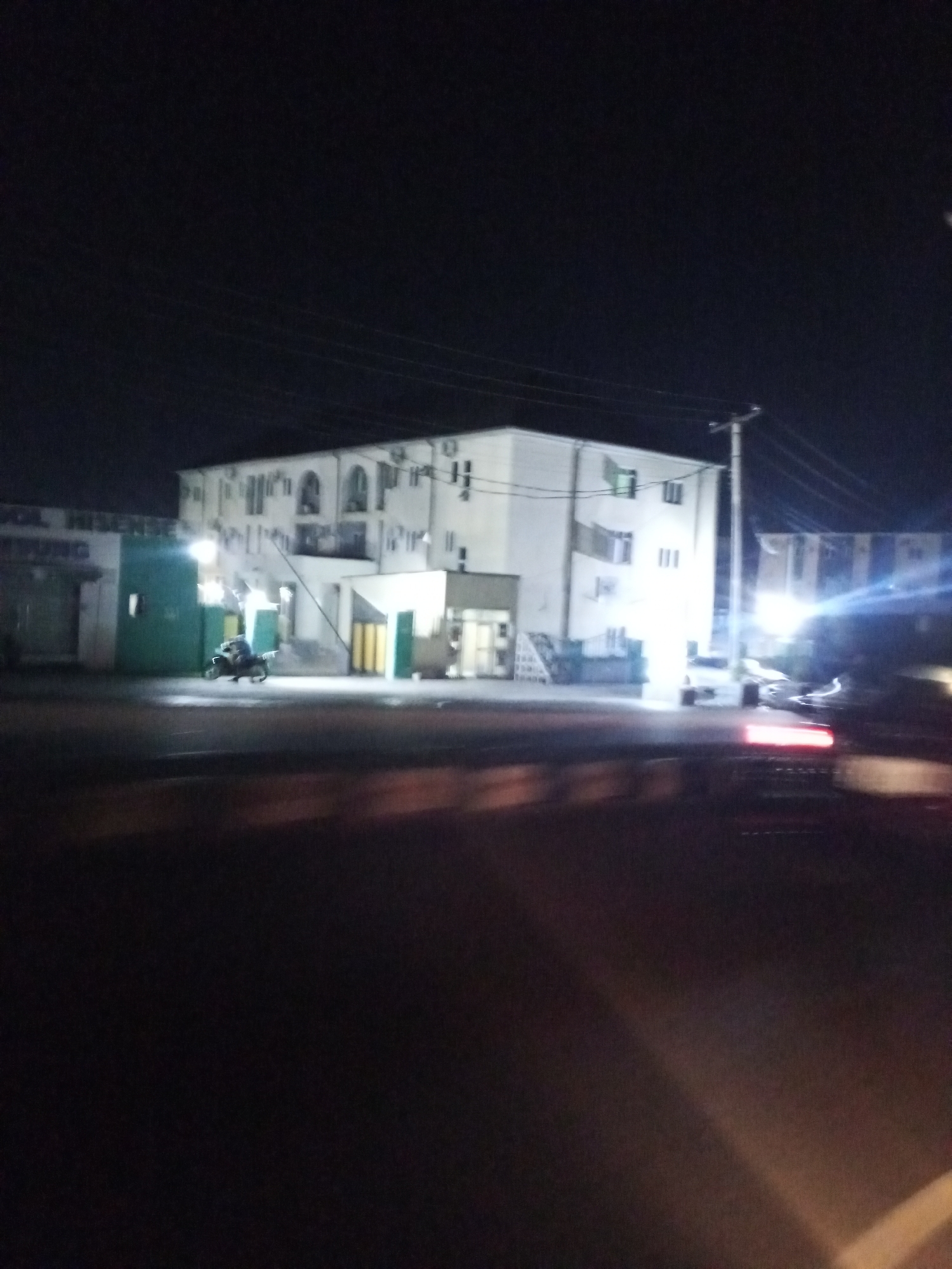 The image captures a nighttime street scene in Lokoja, Nigeria, with a focus on a multi-story building illuminated by artificial lights. The building appears to be a commercial establishment or residence, with several windows visible on its white facade. A motorcycle is parked in front of the building, and a person is seated on it. Adjacent to the building is a green structure, possibly a shop or office, with text visible above it that includes the word "HISENSE". A utility pole with overhead wires stands to the right of the building. The foreground is blurred, suggesting the photo was taken from a moving vehicle, and shows the taillight of another vehicle. The overall impression is of a typical urban night environment.
