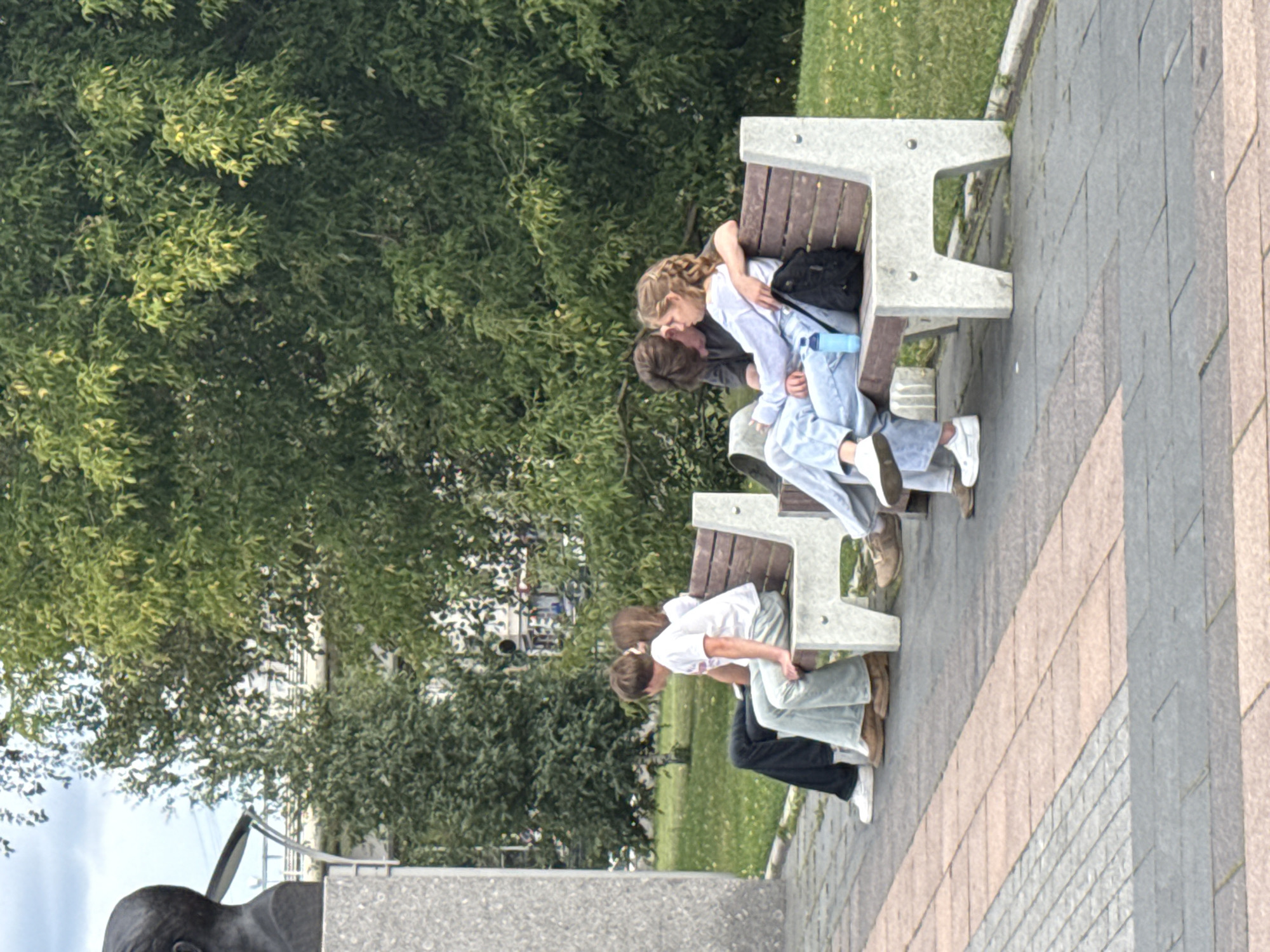 In Moscow, Russia, two couples are seated on a park bench. The couple on the right is in the foreground, with the woman leaning into the man who has his arm around her. She is wearing a light blue sweatshirt and light blue jeans. The man is wearing a dark gray t-shirt. A blue water bottle and a black backpack are on the bench near them. The couple on the left is in the background, sitting further apart. The man is wearing dark pants and a white t-shirt, and the woman is wearing a white t-shirt and light-colored pants. The bench is made of concrete and wood. The background features lush green trees and grass. A stone monument with a bust is visible on the left side of the image, behind the first couple. The ground in front of the bench is paved with alternating patterns of light and dark gray and pinkish-tan tiles. The overall scene suggests a relaxed atmosphere in a public park setting.