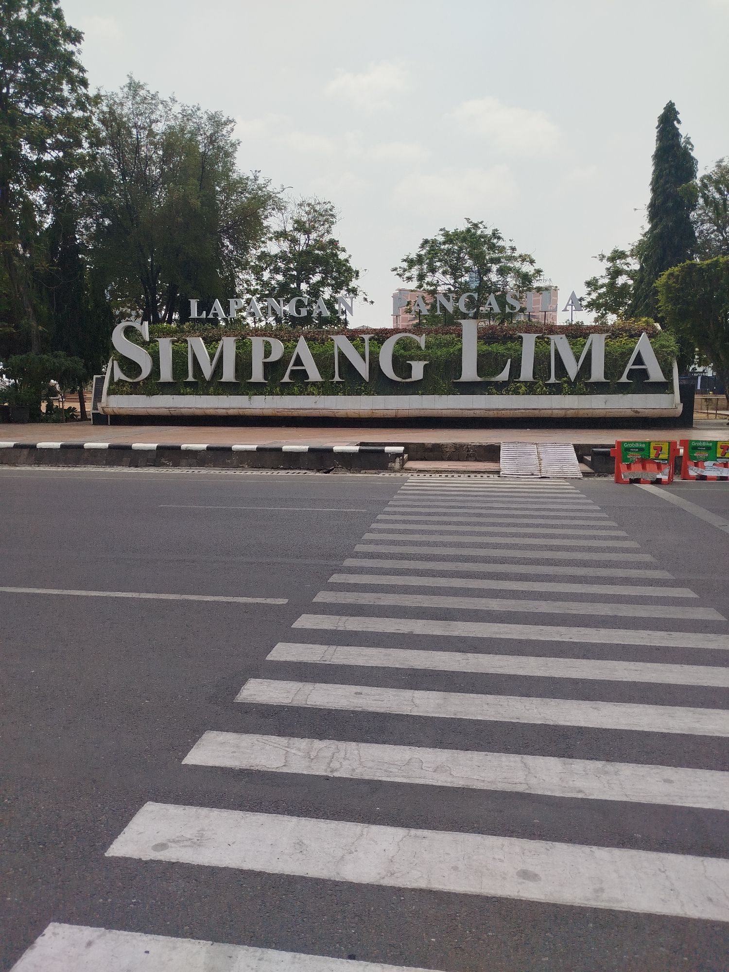 A wide-angle shot captures a city landmark in Semarang, Indonesia, identified by large white letters spelling "LAPANGAN SIMPANG LIMA". The letters are set against a backdrop of lush green bushes and a cloudy sky. In the foreground, a pedestrian crossing with white stripes on asphalt leads towards the landmark. Orange traffic barriers with "GrabBike" advertisements are visible on the right side of the road. The scene appears to be an outdoor public space, likely a busy intersection or a park entrance.