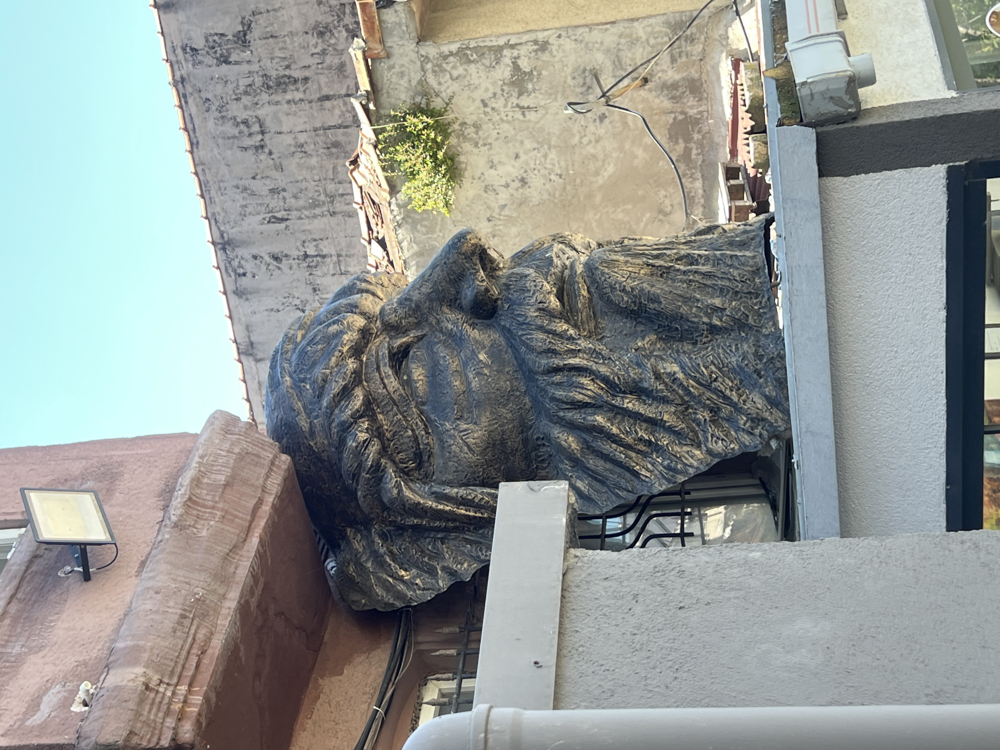 Captured from a low-angle perspective, the image reveals a colossal head sculpture positioned adjacent to a building. The sculpture, rendered in a dark metallic hue, possibly bronze or iron, is highly textured and depicts a stern or contemplative visage. Its features – nose, brow, and hair – are prominently defined.
The buildings flanking the sculpture appear to be weathered, displaying varying architectural styles. The structure to the left is coated in a light reddish-brown stucco or plaster, featuring a window and a mounted outdoor light. The building on the right presents a modern facade with white textured walls and dark-framed windows. Overhead, a light blue sky contrasts with the aged structures and sculpture.
This image was taken in Şişli, Turkey, where this sculpture is a local landmark.