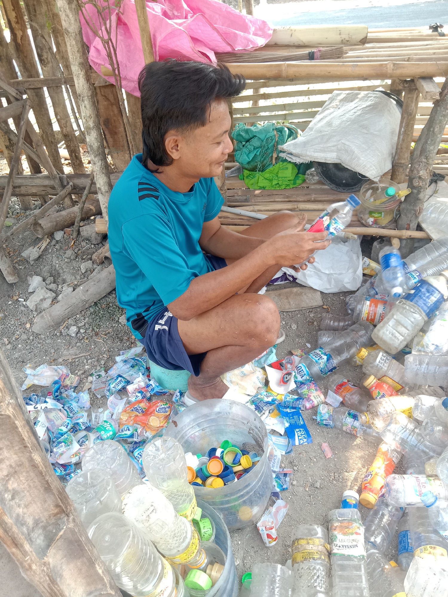 A man, wearing a teal t-shirt with black stripes on the sleeves and dark blue shorts, is sitting on the ground in a makeshift shelter constructed of bamboo poles and planks. He is surrounded by a large collection of plastic bottles, bottle caps, and various wrappers. The man is holding a clear plastic bottle in his hands, appearing to examine it. The ground is covered in dirt, small rocks, and debris. In the background, there are more plastic items, a large white bag, and a pink tarp. The scene suggests a location where recyclables are being collected or sorted, potentially in a roadside area of Stiaji, Indonesia.