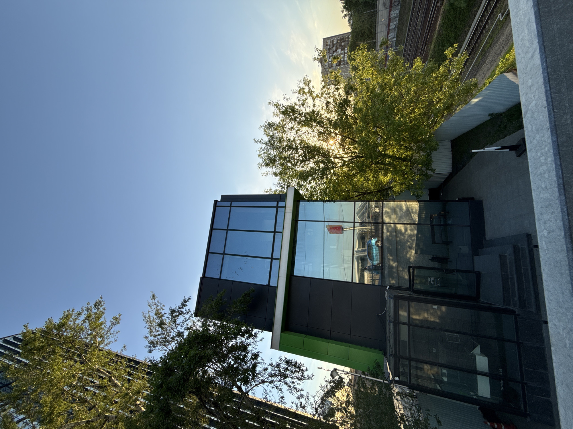 The image shows a modern building with a glass facade located in Moscow, Russia. The building has a black and green exterior with large windows reflecting the surrounding environment. A tree with green leaves partially obscures the building. In the background, a railway line and a taller building can be seen. The sky is blue with a few clouds, and the sun appears to be shining through the tree branches. The image appears to be taken from an elevated perspective. There are a few people in the reflection of the windows.