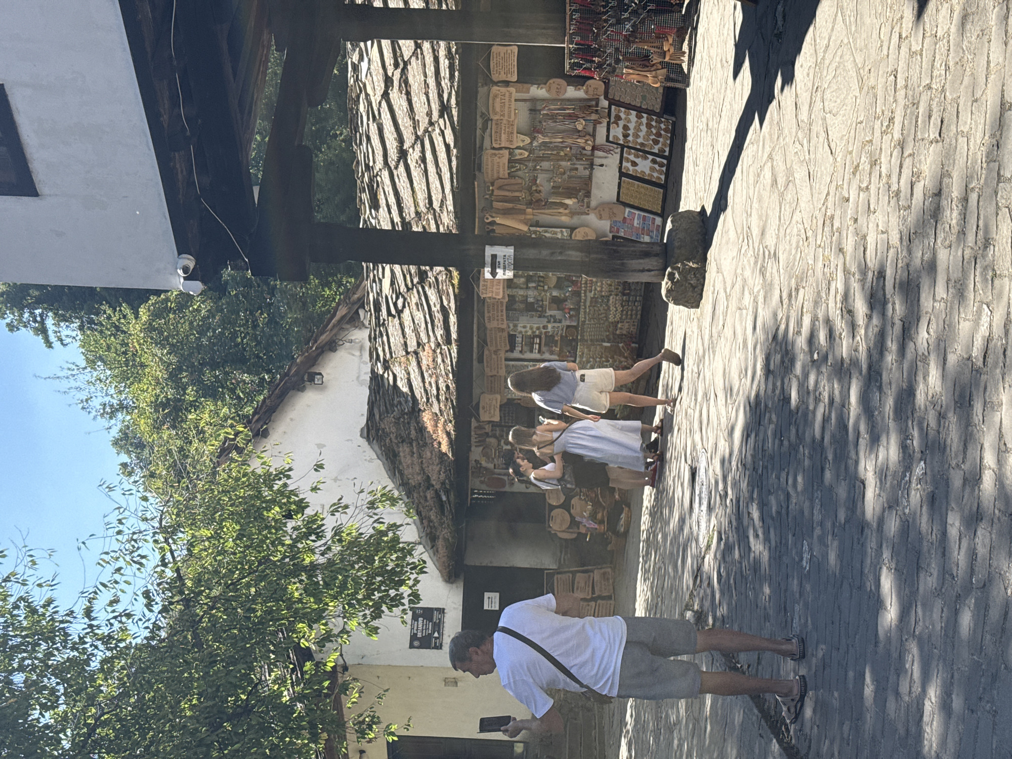 The image depicts an outdoor scene in the city of Dryanovo, Bulgaria, likely in an area with traditional architecture and artisan shops. In the foreground, a man wearing a white t-shirt and grey shorts is holding up a mobile phone, possibly taking a picture or video. He is facing away from the camera, and a dark strap from a bag or satchel is visible across his chest.
Behind him, a group of three women are standing near a shop. One woman in a white dress, another in a dark top and skirt, and a third in a light blue top and white shorts. They appear to be browsing or interacting with the shop's offerings.
The shop itself is characterized by a rustic, tiled roof and wooden beams. It appears to be selling various handcrafted items, such as wooden utensils, keychains, and other souvenirs displayed on racks and tables. Several signs with text in Cyrillic are visible on the shop's facade.
The ground is paved with cobblestones, and patches of sunlight and shadow create a dappled effect. Lush green trees and foliage frame the scene, suggesting a pleasant, sunny day. In the upper right of the image, a white building with a dark wooden overhang is visible, and a security camera is mounted on the overhang. A sign on the building in the background reads "DRYANOVO."