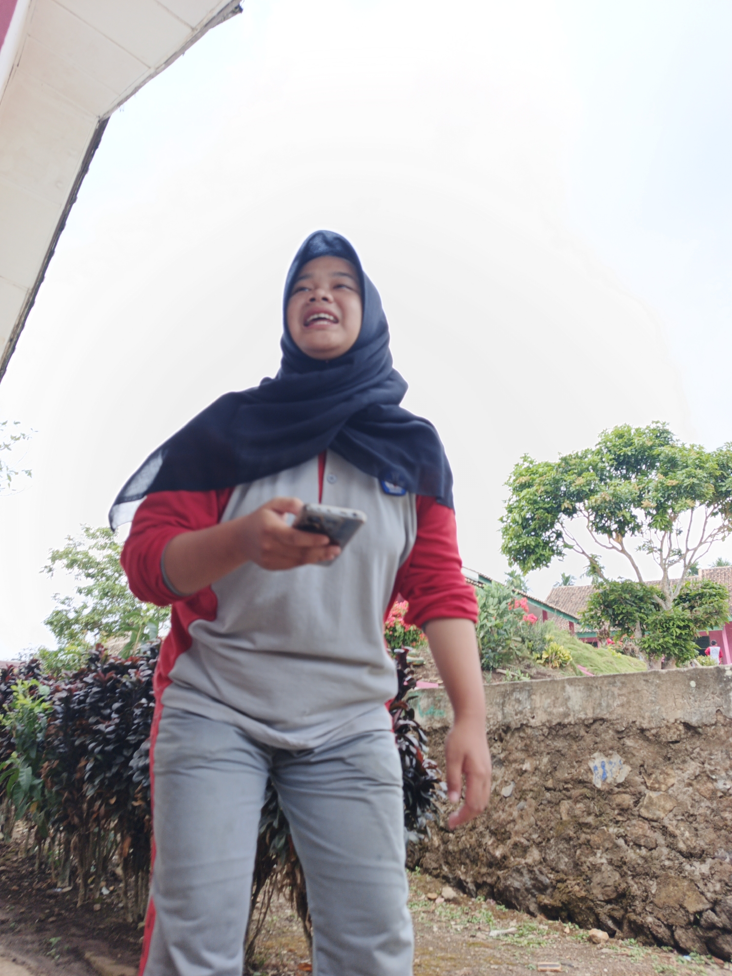 A young woman wearing a navy blue hijab and a grey and red athletic outfit stands outdoors, holding a mobile phone. She is smiling widely and appears to be looking upwards. The background features a wall made of rough stones, a lush green tree, and some buildings with tiled roofs, suggesting an outdoor school or community setting. The overall scene is bright and appears to be during the daytime. The location is identified as Tanjung Setia, Indonesia.