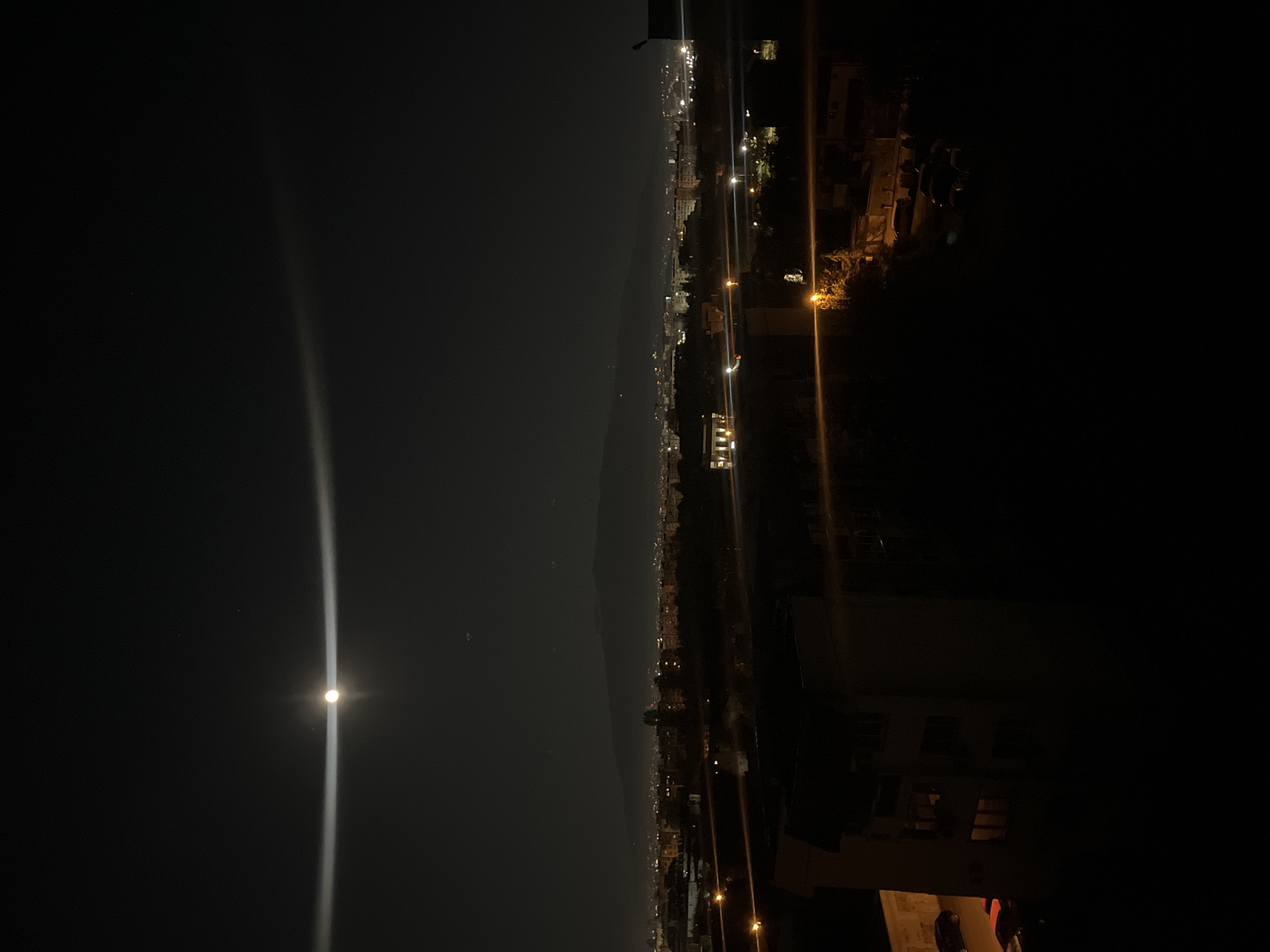 The image shows a night scene, possibly in or near the city of Sofia, Bulgaria. The left side features a dark sky dominated by a bright moon and its reflection or glare extending toward the top. The city is visible as a cluster of lights stretching across the middle ground, separated from the dark sky by a vague line of elevated terrain, potentially mountains. The right side shows urban structures, including buildings illuminated by streetlights or interior lights, adding to the cityscape. The image appears to be taken from an elevated position, offering a broad view of the city at night.