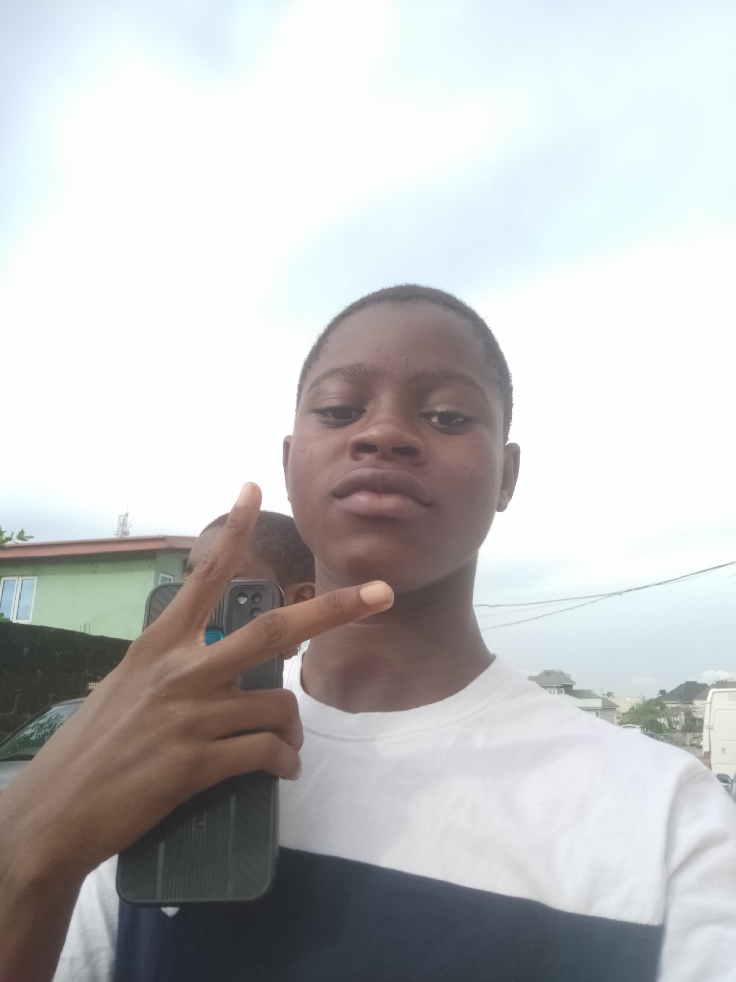 The image is a waist-up shot of a young person posing outdoors. The subject, who appears to be male with dark skin and short dark hair, looks directly at the camera with a neutral expression. They are wearing a short-sleeved t-shirt that is predominantly white, with a wide, dark blue or black horizontal stripe across the lower chest.
The subject's left hand is raised, forming a "peace sign" (V-sign) with the index and middle fingers extended. They are holding a black smartphone, encased in a textured dark cover, in front of their torso. Part of another person's head, also with dark skin and short hair, is faintly visible behind the main subject's raised hand, positioned slightly to the left.
The background shows an outdoor setting under a bright, overcast sky, indicating daytime. To the left, a light green building with white-framed windows is visible behind a dark wall or fence. A vehicle is partially obscured by the fence in the lower left corner. Faint power lines or cables stretch across the upper right portion of the background. The lighting is soft and diffused, creating few harsh shadows. There is no visible text in the image.