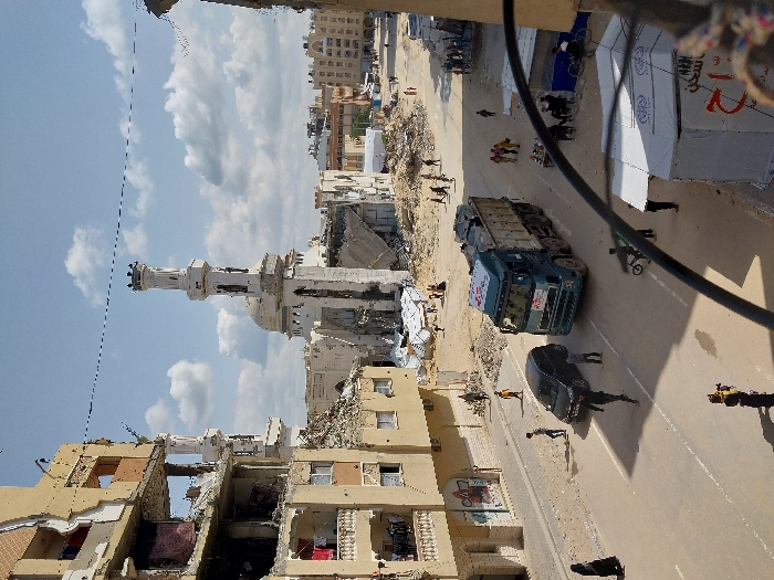 This image depicts a street scene in Khan Yunis, Palestinian Territory, under a partially cloudy sky. The environment shows extensive damage consistent with conflict. In the background, a large mosque with a prominent white minaret and dome is severely damaged, with significant structural collapse and surrounding rubble. Multi-story buildings line both sides of the asphalt street, exhibiting considerable destruction, including missing walls, exposed interiors, and debris; laundry is visible hanging from balconies on the left building.
On the street, a blue dump truck, bearing a Red Crescent emblem on its cab, is moving towards the viewer, loaded with what appears to be construction debris. A black passenger vehicle is positioned on the left side of the road. Numerous pedestrians are present: walking on the street, near the damaged buildings, and amidst the rubble around the mosque. A person holding a camera or recording device is visible in the lower left foreground, and a child on a bicycle is near the dump truck. On the right side, two white temporary shelters or aid tents, one displaying the World Food Programme (WFP) logo and Arabic text, are erected near the roadside. A dark, curved object, likely a cable or railing, partially obstructs the immediate foreground.