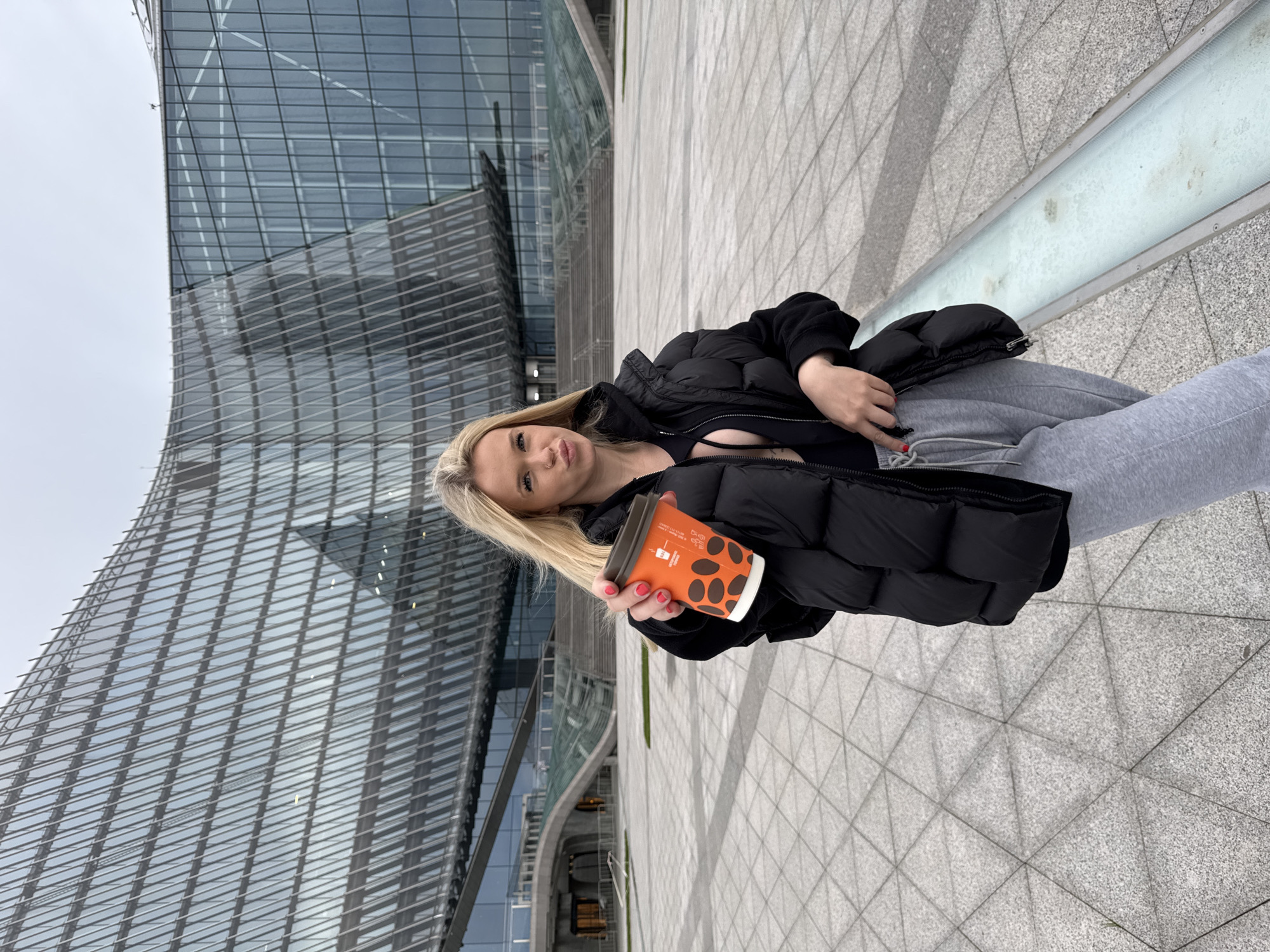 This media file shows a young blonde woman holding a coffee cup and standing in front of a modern building with a glass facade. She is wearing a black puffer jacket, a black top, and gray sweatpants. The coffee cup is orange with black polka dots and has some text on it that is not clearly legible. The woman is looking at the camera with a playful expression. The background is a paved area with a geometric pattern and a cloudy sky. The building appears to be made of glass and metal, with a curved design. The scene suggests an urban setting in Saint Petersburg, Russia. The lighting indicates it could be daytime, possibly overcast due to the cloudy sky. The woman's attire and the coffee cup suggest a casual outing.