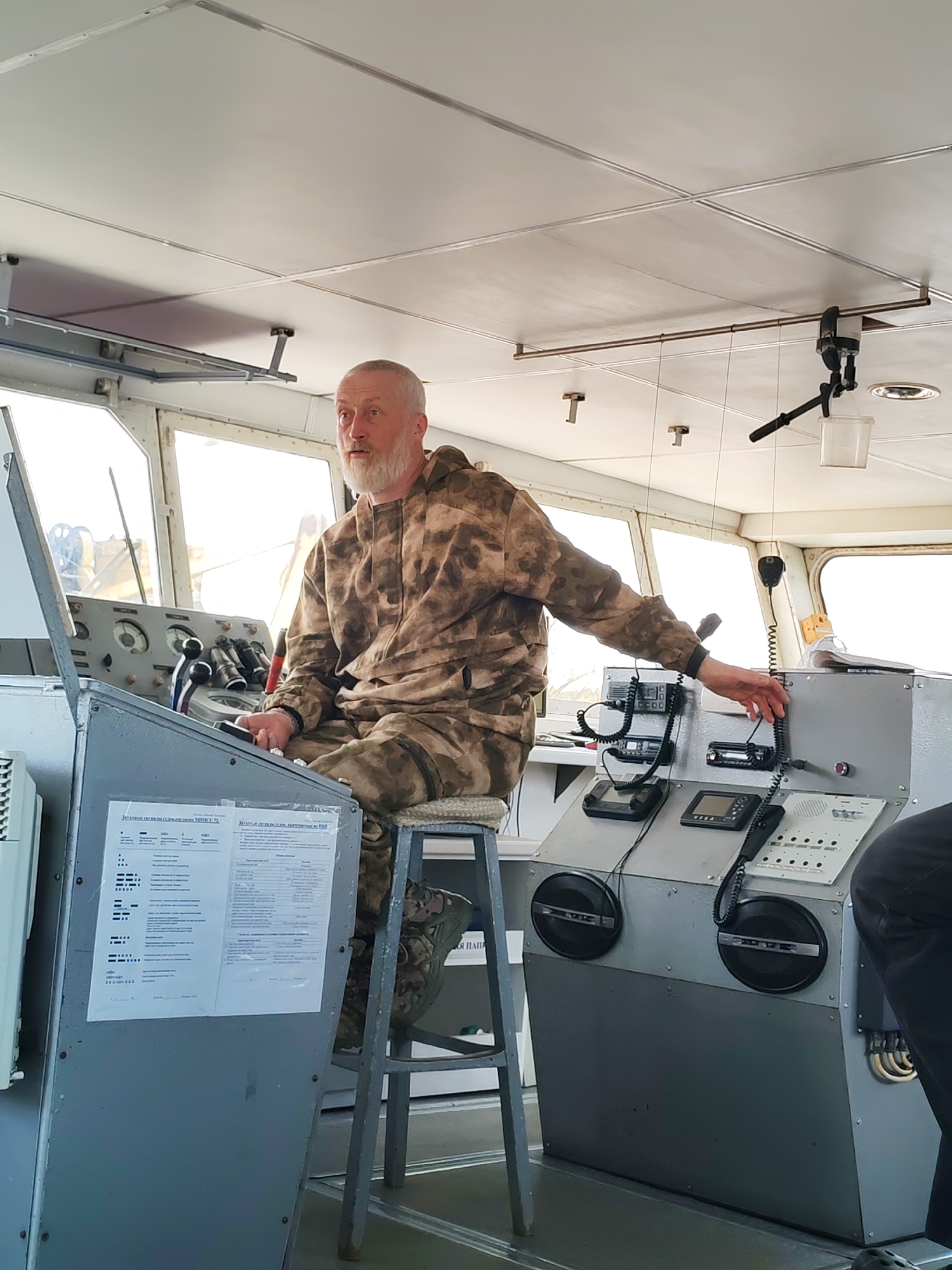 A man with a gray beard and wearing camouflage clothing is seated on a high stool inside the cabin of a ship. He is positioned at a control console with various instruments and communication devices. The interior is utilitarian, with gray metal panels and a white ceiling. Daylight streams through the windows, suggesting an outdoor location.
Although the location of Yakhroma, Russia, is mentioned, there is no visual information in the image that explicitly confirms the location.