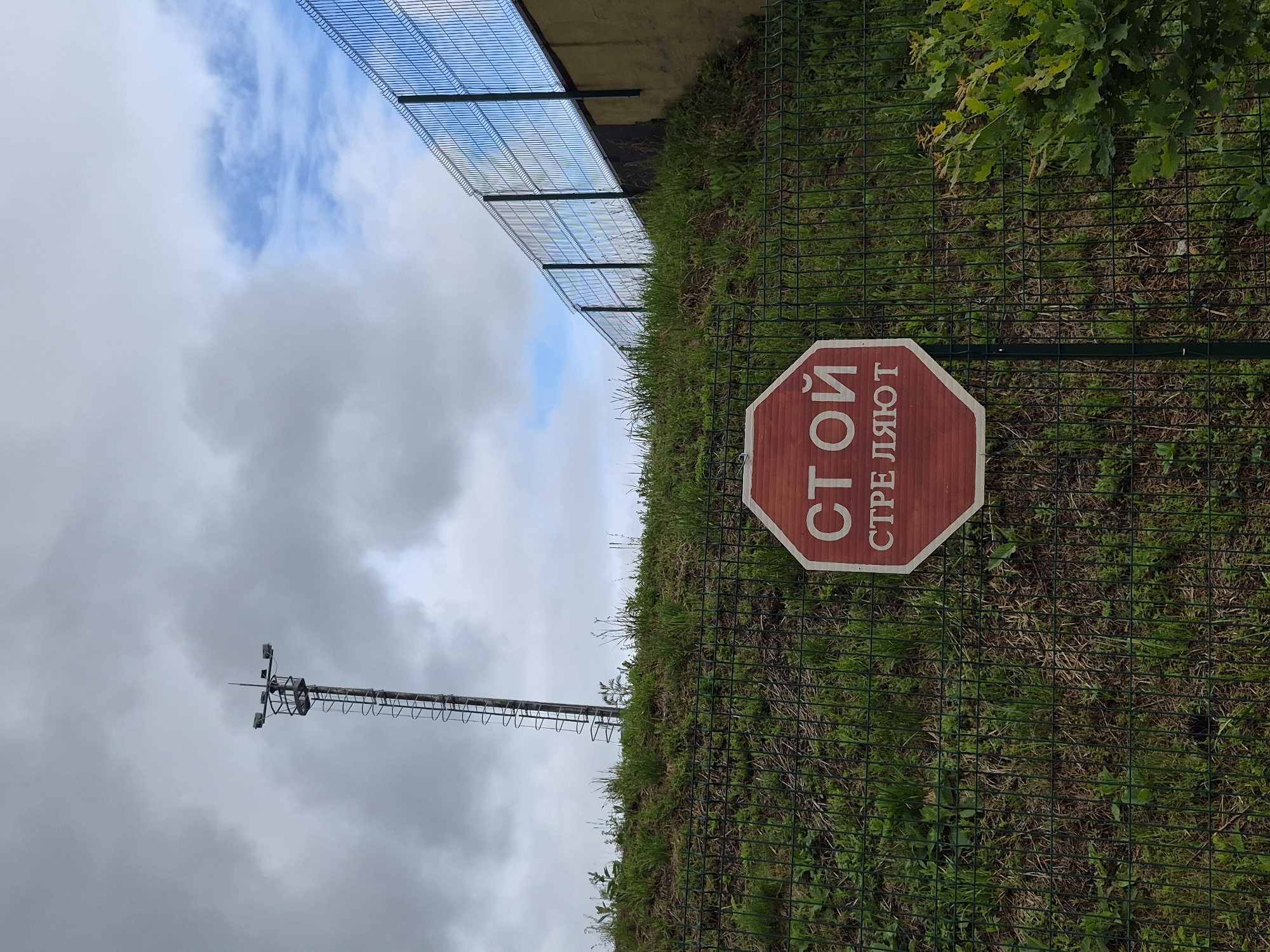 An outdoor scene captured under a partly cloudy sky, with patches of blue visible in the upper right quadrant. In the upper left, a tall metal tower with an integrated ladder ascends, supporting a platform equipped with multiple surveillance cameras or sensor devices. A green metal mesh fence with vertical posts runs horizontally across the midground, separating the foreground from a grassy embankment. The embankment is covered in green grass and various small plants. To the right, the fence continues, running alongside a yellowish-brown wall or structure. In the lower-center foreground, attached to a fence post, is an octagonal red sign with white Cyrillic text. The top line reads "СТОЙ" (STOY), translating to "STOP." The bottom line reads "СТРЕ ЛЯЮТ" (STRE LYAYUT), translating to "SHOOTING" or "THEY ARE SHOOTING." A green leafy bush is visible in the lower right corner, partially obscured by the fence. No individuals are present. This scene is located in Прикамский, Russia.