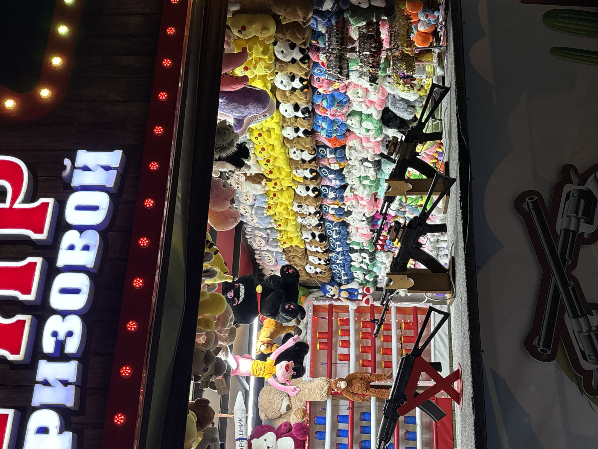 The image shows a close-up view of a carnival game booth at night, likely at an amusement park or fair. The booth features a large, illuminated sign at the top with Russian text in red and white letters, reading "ПРИЗОВОЙ" (meaning "Prize"). Below the sign, a row of red LED lights adds to the festive atmosphere.
The main focus of the booth is the display of numerous plush toys, primarily stuffed animals of various shapes and colors, arranged on shelves and hanging from the ceiling. These include teddy bears, characters resembling Pikachu and Sonic the Hedgehog, and many other generic animal figures. In front of the plush toys, there are several toy rifles mounted on stands, suggesting a shooting gallery game where participants aim at targets (presumably some of the plush toys) to win prizes. Some of the rifles appear to be assault rifle replicas.
The setting appears to be an outdoor or semi-outdoor venue, given the lighting and the general ambiance. The presence of the specific Russian text on the sign indicates a location in Russia. While the exact location within Novorossiysk is not specified in the image itself, it is plausible that this image was taken at a fairground or entertainment complex within the city of Novorossiysk, Russia, during a festive event or regular operation. There are no people visible in the image.