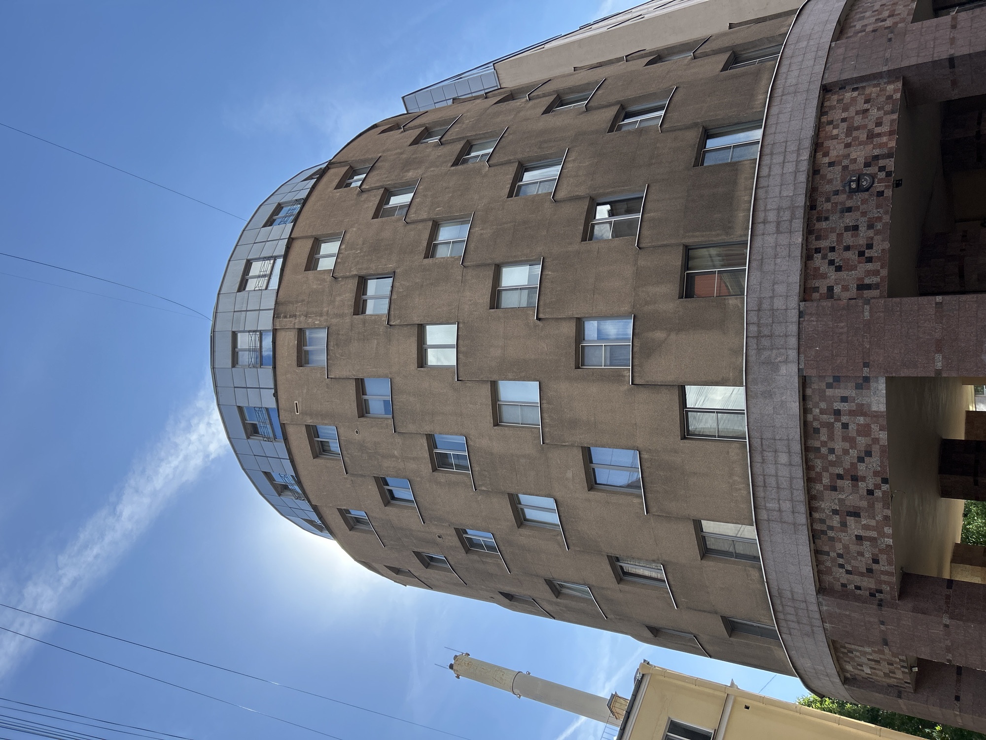 A tall, multi-story building with a distinctive curved facade is viewed from a low angle against a clear blue sky. The building's main body is clad in a textured, dark beige or grey concrete, featuring numerous irregularly placed rectangular windows, some with sloped sills. One side of the building presents a smooth, reflective glass curtain wall that follows the curve, contrasting with the rough concrete texture.
On another side, a decorative mosaic-like pattern of brown, red, and black tiles adorns a section of the facade, separated by a wide, curved band of grey material. A small, dark fixture is visible on the tiled area. Below the main building, a tall, slender, light-colored chimney-like structure rises, and a portion of a yellow building is visible. Overhead, several power lines or cables stretch across the expansive blue sky.
The scene is set during bright daylight hours, indicated by the clear sky and sunlight, with a few faint wispy clouds or contrails present. No people or specific activities are depicted. No visible text is present in the image.