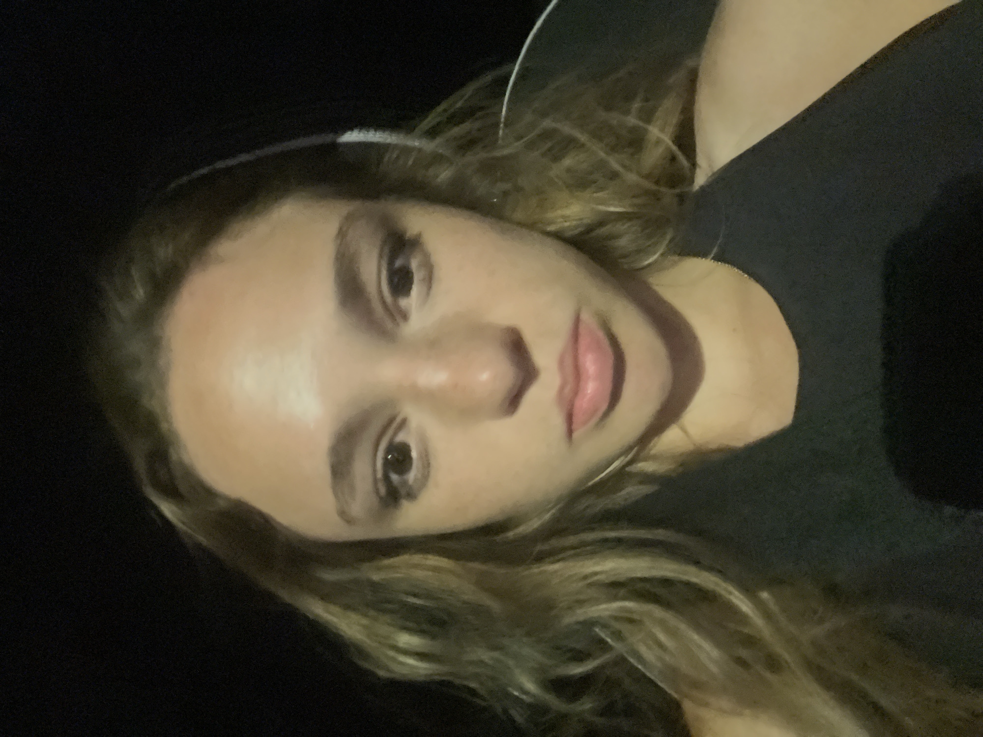 A close-up selfie of a fair-skinned young woman with light brown hair. She is wearing a black t-shirt. A white cable, possibly from headphones, is visible near her hair. The background is dark and indistinct. The location is not discernible from the image.