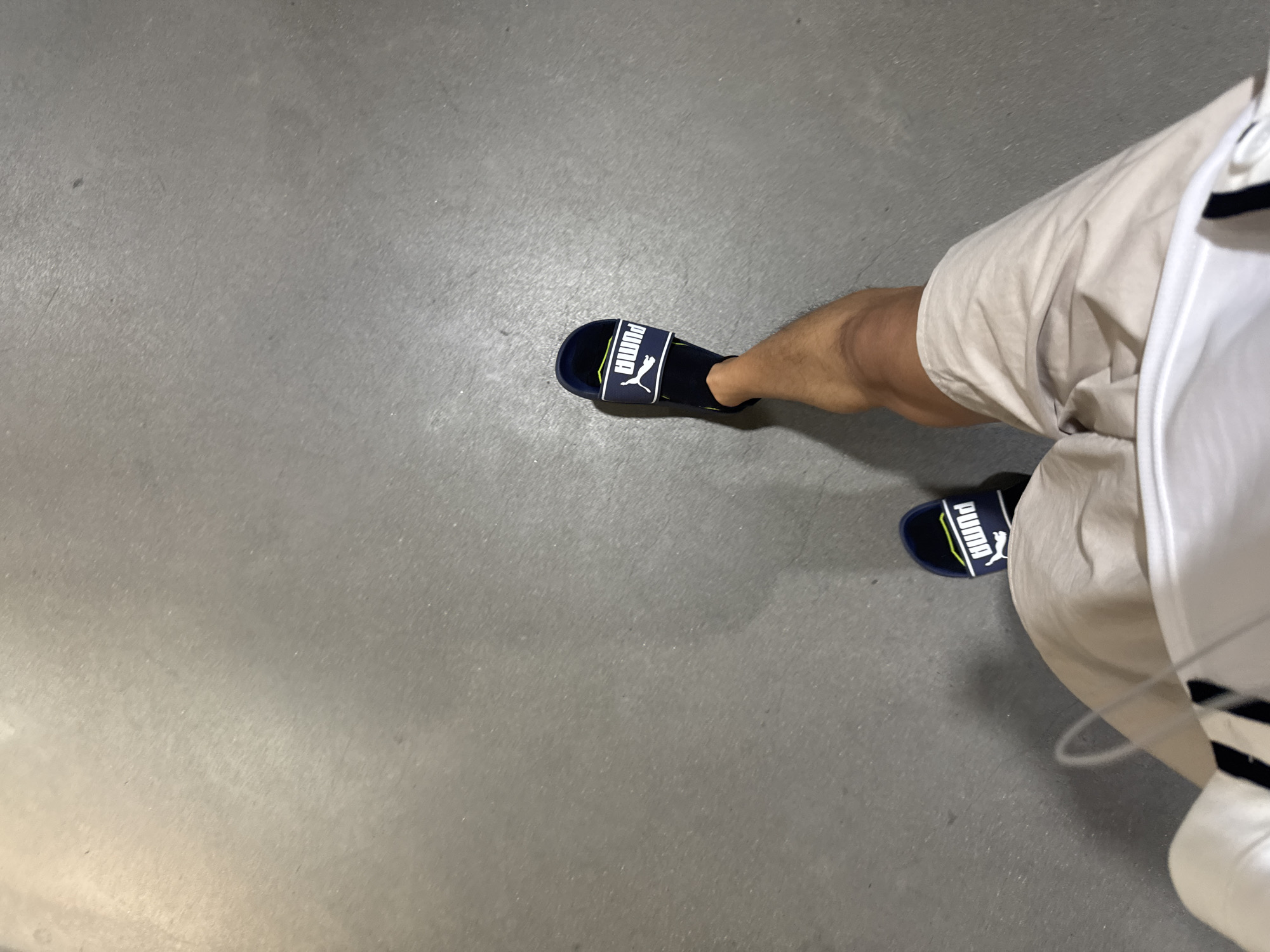 The image displays an overhead, first-person perspective, looking down at a person's lower body on a uniform grey surface. The surface appears to be concrete or a similar paved material, showing some texture and minor irregularities.
The subject is wearing dark blue (or black) Puma slides on their feet. Each slide features the brand name "PUMA" in white capital letters across the strap, accompanied by the recognizable white leaping puma logo. A hint of lime green or yellow is visible on the inner sole of the slides.
The person's legs are visible, and they are dressed in light-colored, possibly beige or khaki, shorts. A portion of a white top with a dark striped detail on the collar or sleeve is visible on the right side of the frame, along with a thin white wire, likely from headphones.
The lighting is soft and even, without harsh shadows, suggesting an indoor environment or an overcast day. There are no other discernible objects or features in the immediate background beyond the floor. The image captures a casual, everyday scene.